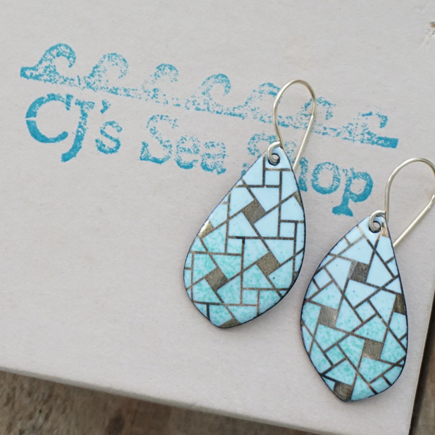 A pair of leaf-shaped dangle earrings with a light green and gold mosaic tile pattern and gold fishhook wires, displayed on a gift box stamped with 'CJ's Sea Shop'.
