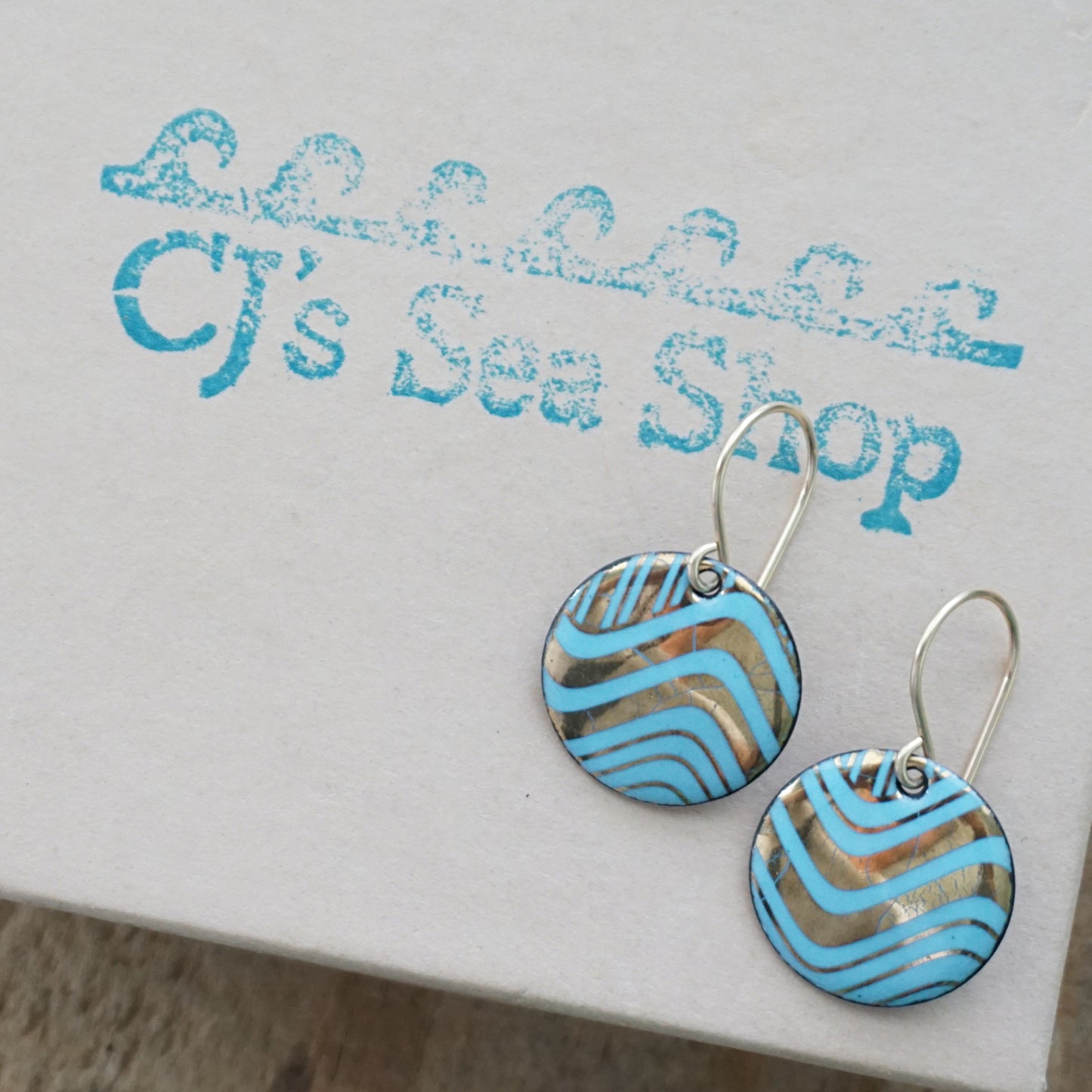 A pair of round drop earrings with light blue and gold wavy patterned enamel and gold-tone fishhook wires, shown on a gift box
