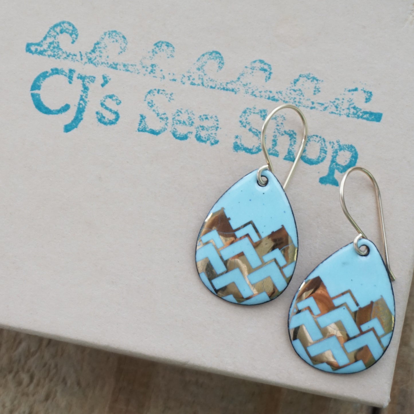 A pair of teardrop-shaped dangle earrings with a light blue enamel background, featuring a metallic gold chevron pattern, hanging from thin gold hooks, displayed on a gift box