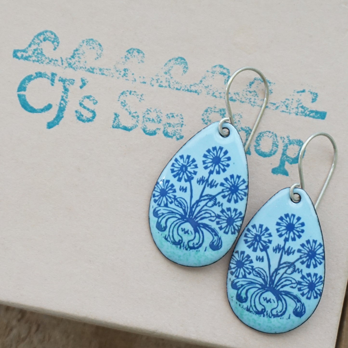 A pair of light blue teardrop earrings with blue floral dandelion designs, displayed on a gift box
