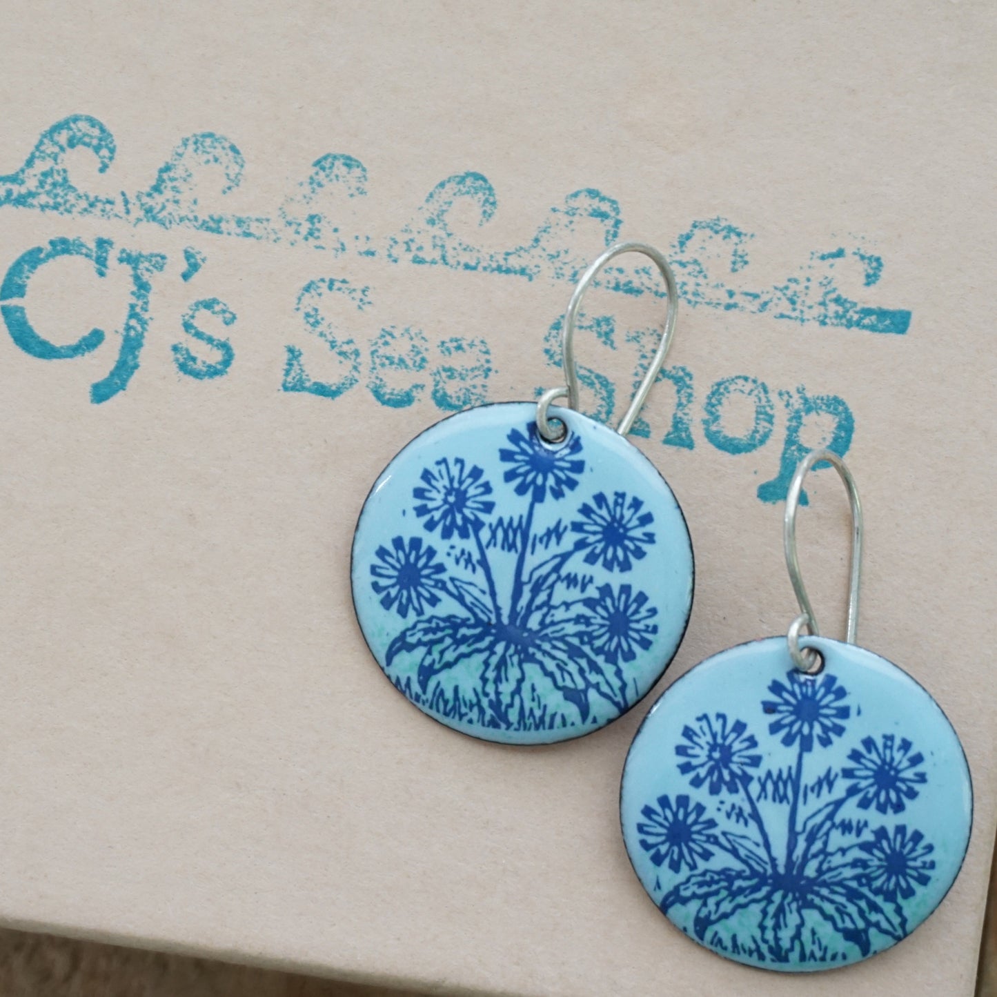 A pair of light blue round earrings with blue floral dandelion designs, displayed on a gift box