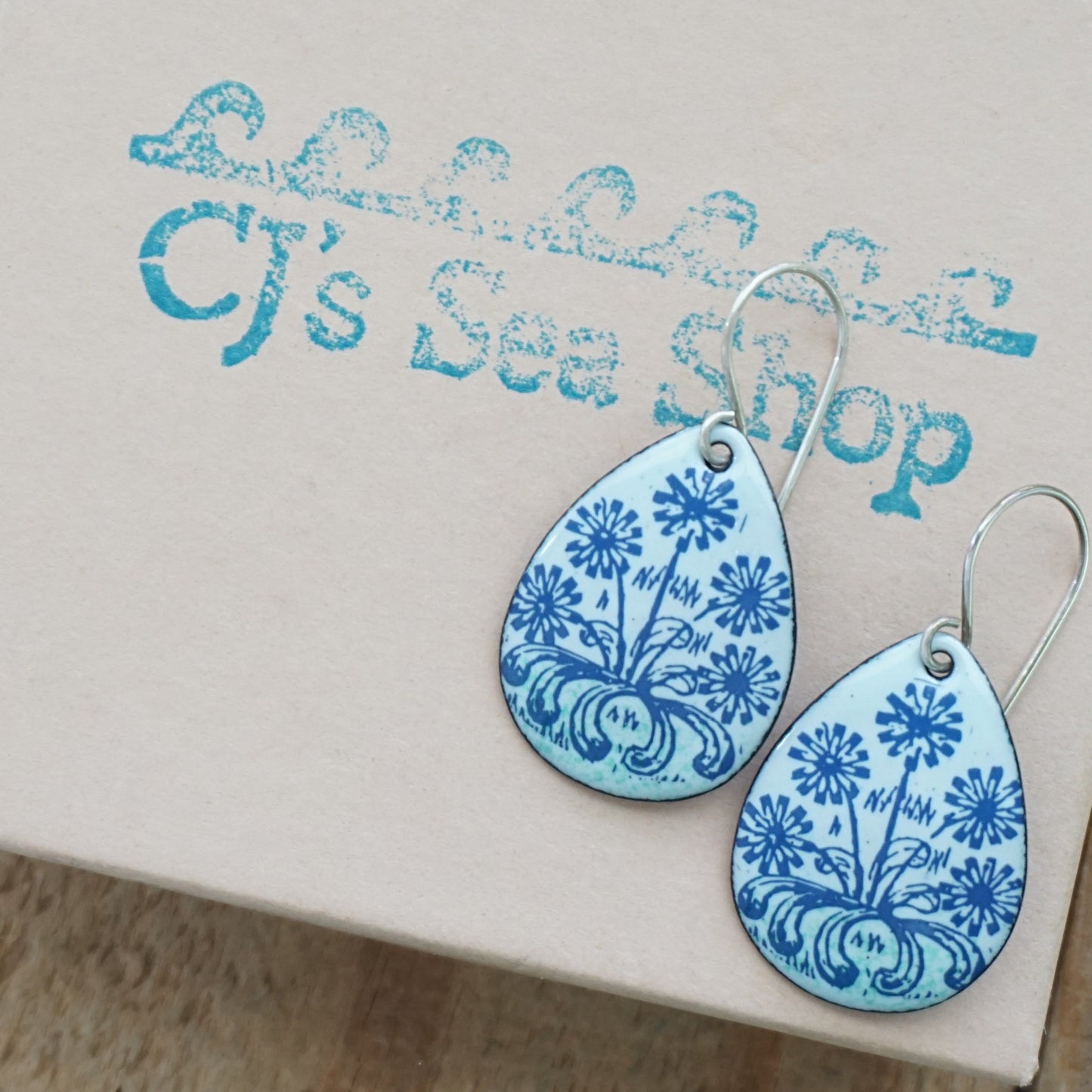 A pair of light seafoam green teardrop earrings with blue floral dandelion designs, displayed on a gift box
