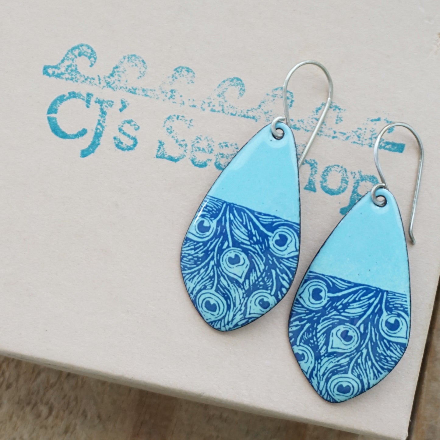 A pair of teardrop-shaped earrings with a light blue top half and a blue patterned bottom half, displayed on a gift box
