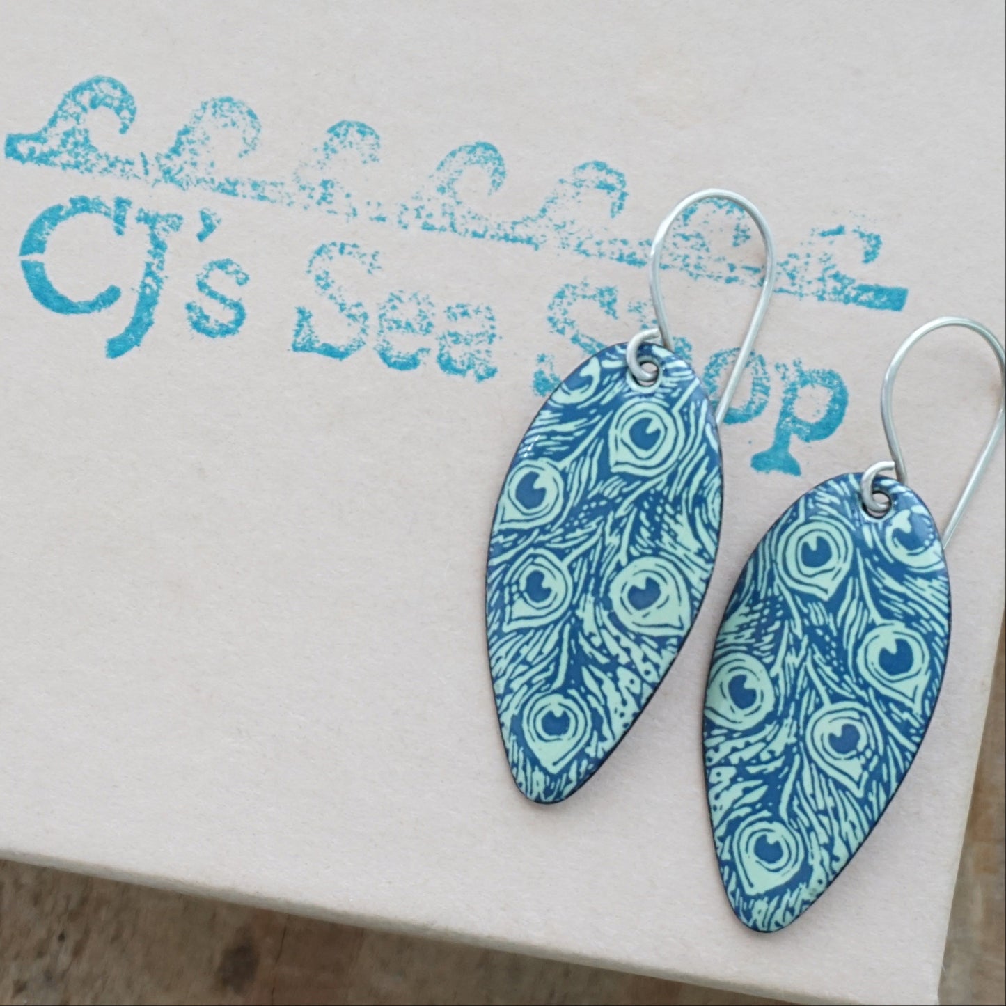 A pair of teardrop-shaped dangle earrings with a blue and light green peacock feather pattern, displayed on a gift box