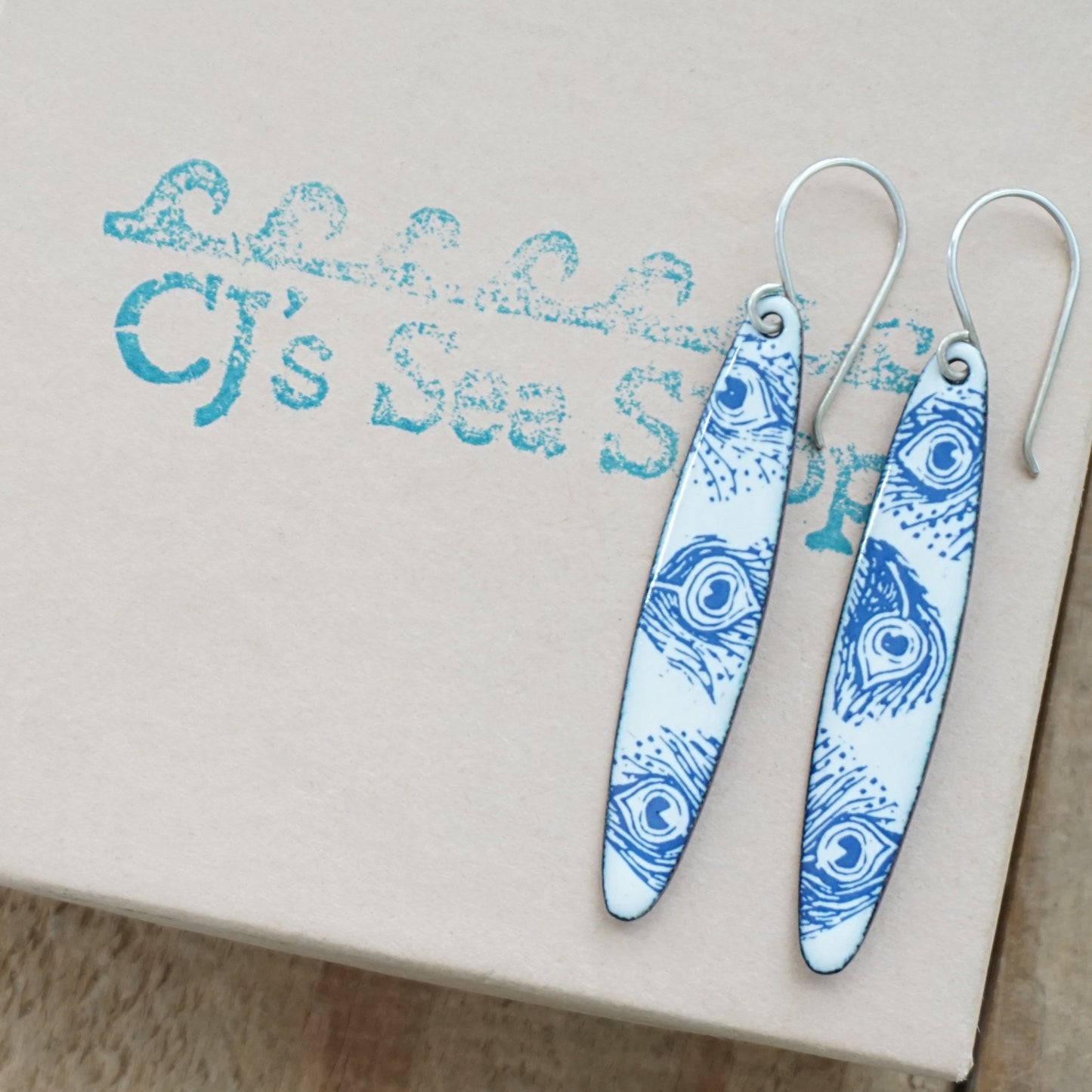 A pair of silver-tone dangle earrings with white enamel inserts printed in blue with a peacock feather pattern, resting on a gift card