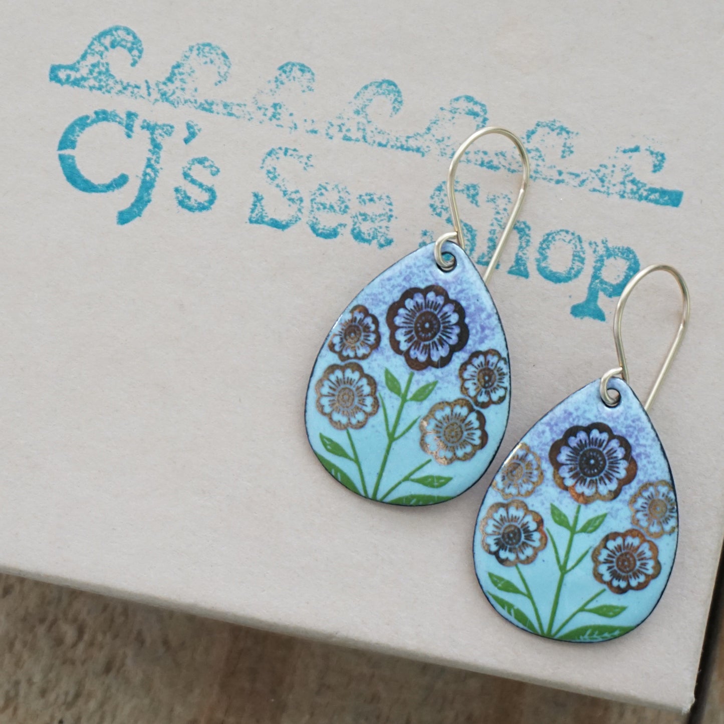 Light blue and purple teardrop-shaped dangle earrings, with gold flowers on green stems dangling from gold hooks, displayed on a gift box