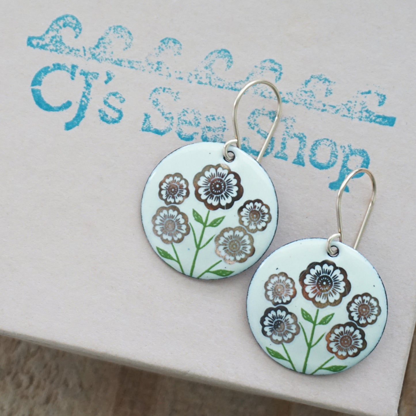 Light yellow disc-shaped dangle earrings, with gold flowers on green stems dangling from gold hooks, displayed on a gift box