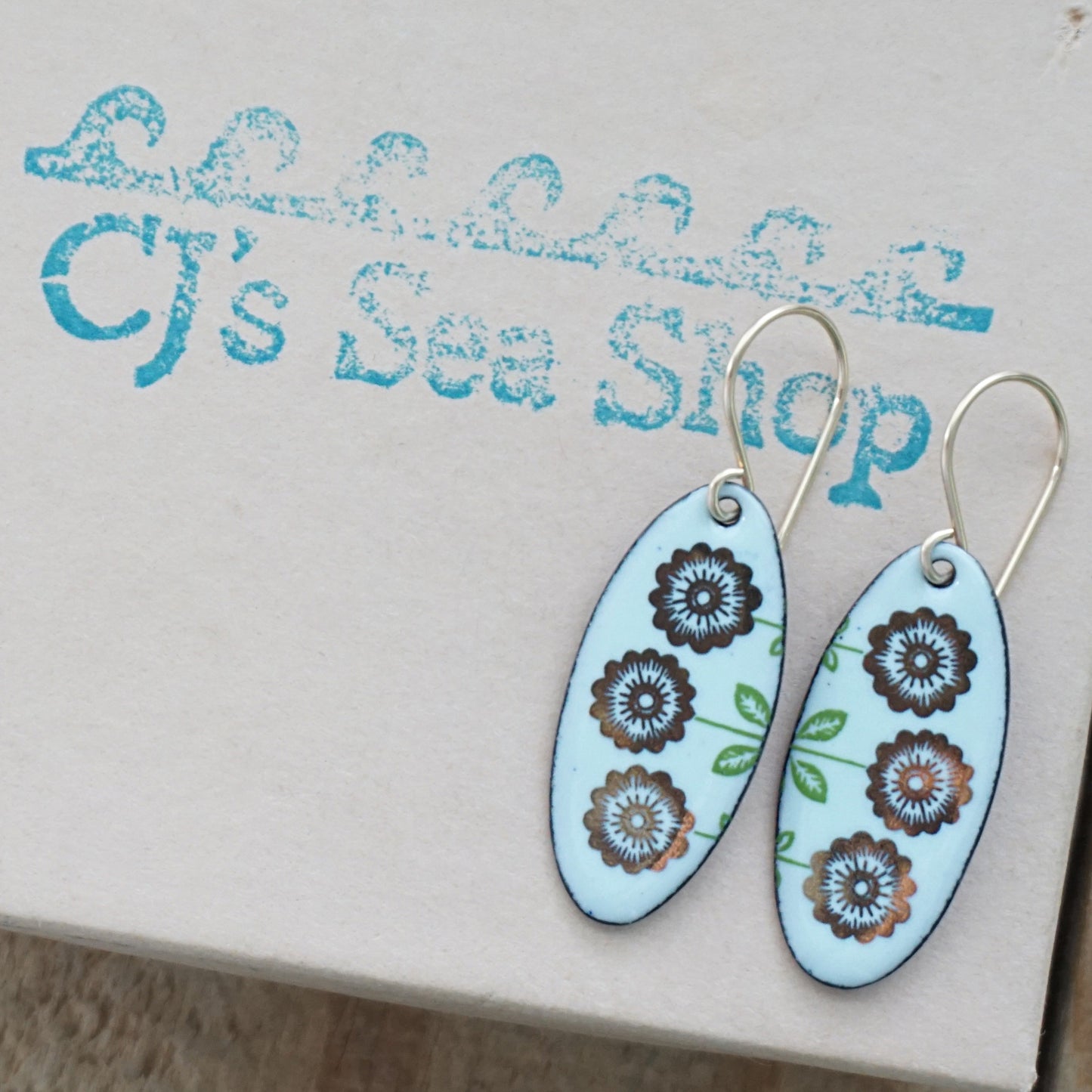 A pair of oval-shaped drop earrings with a light seafoam green background, decorated with gold flowers on green stems, hanging from gold hooks, displayed on a gift box