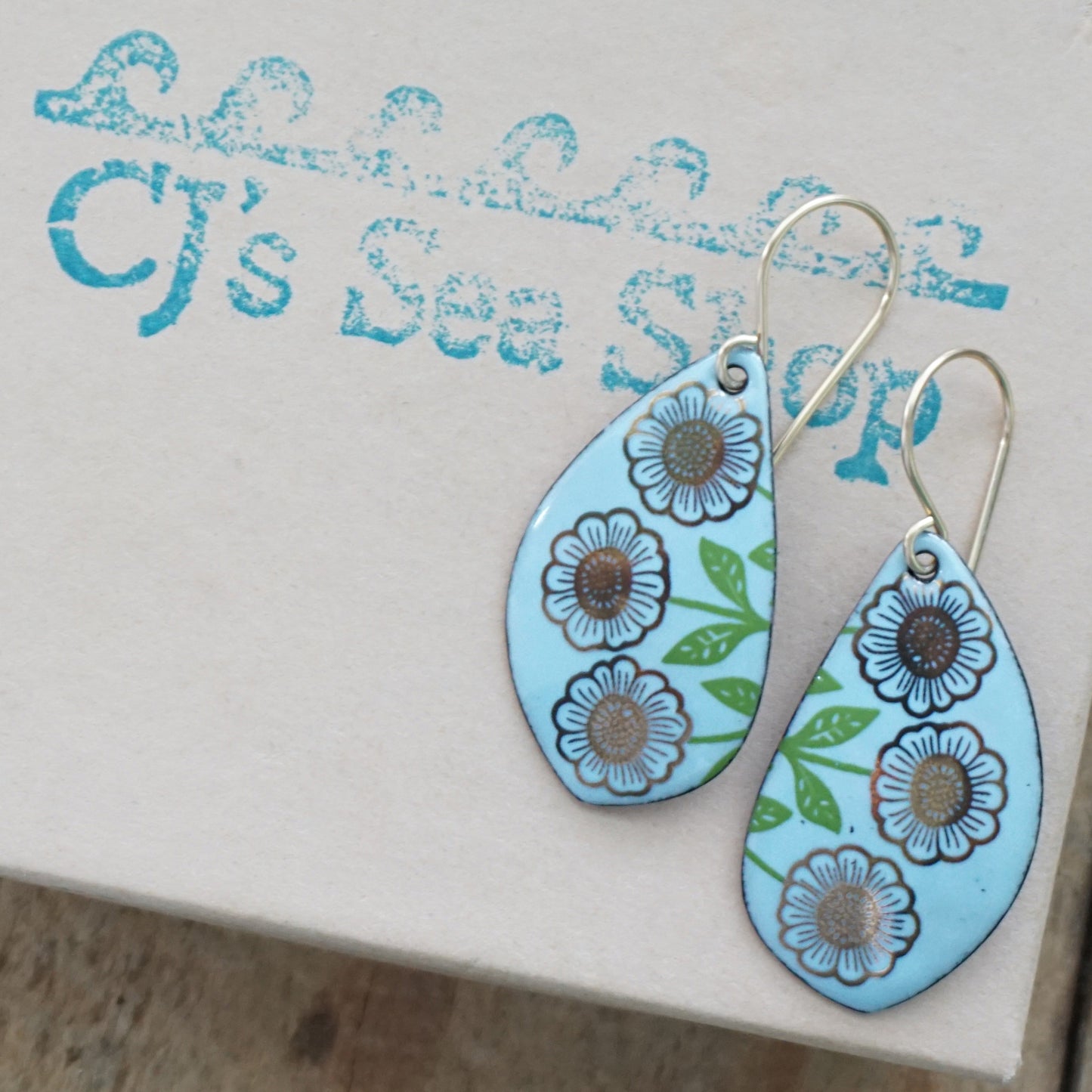 A pair of leaf-shaped drop earrings with a light blue background, decorated with gold flowers on green stems, hanging from gold hooks, displayed on a gift box