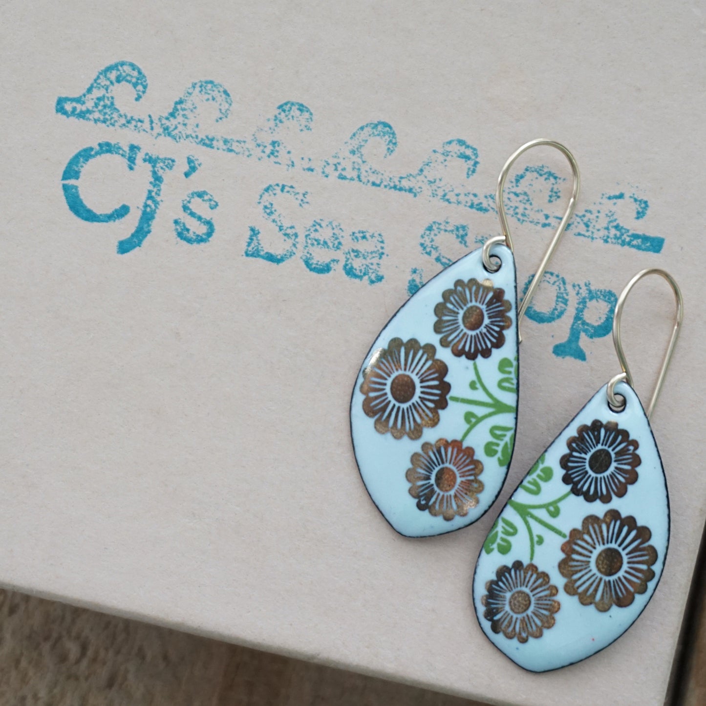A pair of leaf-shaped drop earrings with a light seafoam green background, decorated with gold flowers on green stems, hanging from gold hooks, displayed on a gift box