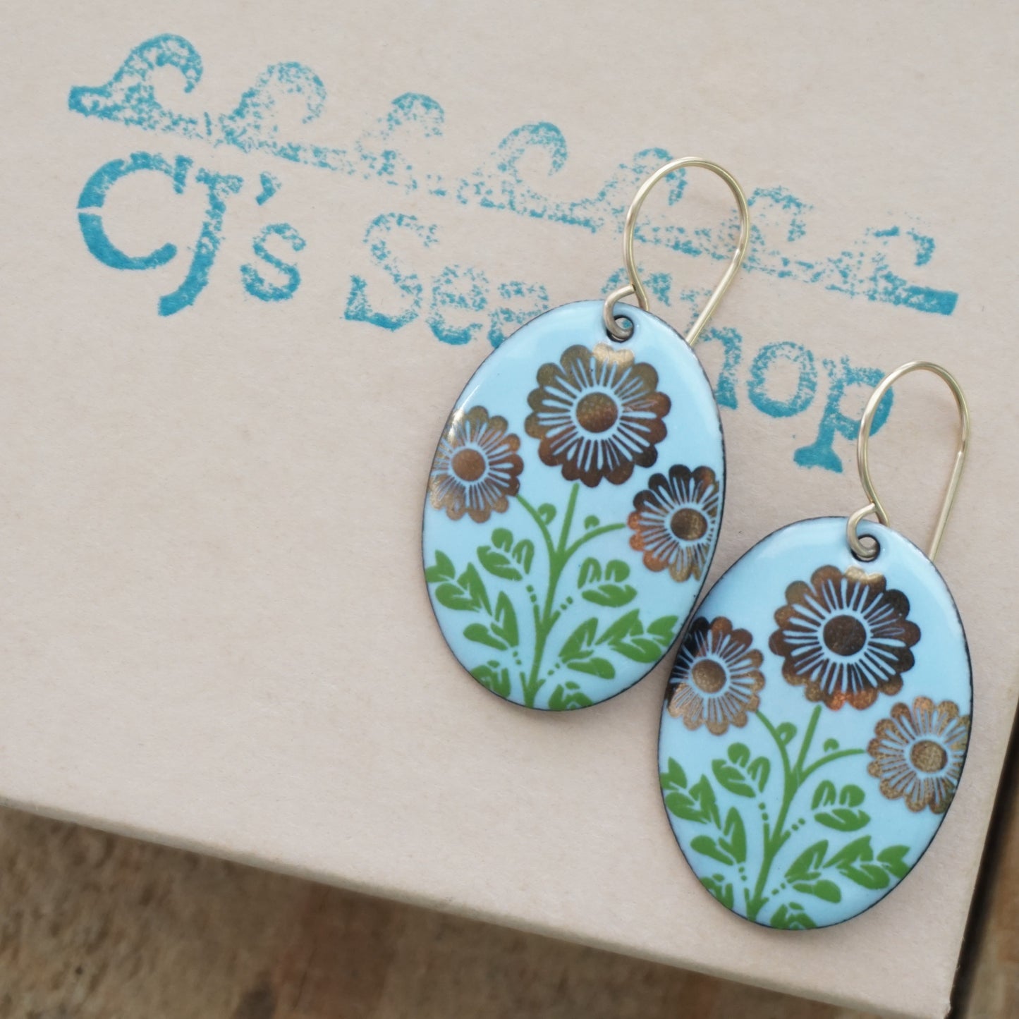 A pair of oval drop earrings with a light blue background, decorated with gold flowers on green stems, hanging from gold hooks, displayed on a gift box