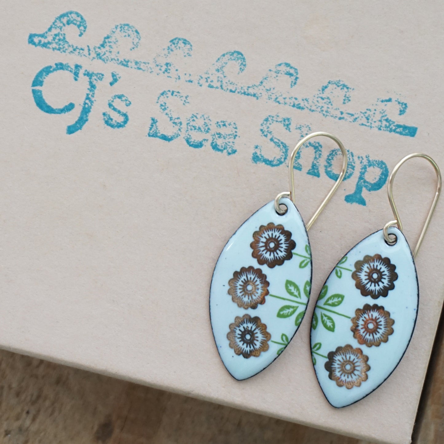 A pair of marquis-shaped drop earrings with a light seafoam green background, decorated with gold flowers on green stems, hanging from gold hooks, displayed on a gift box