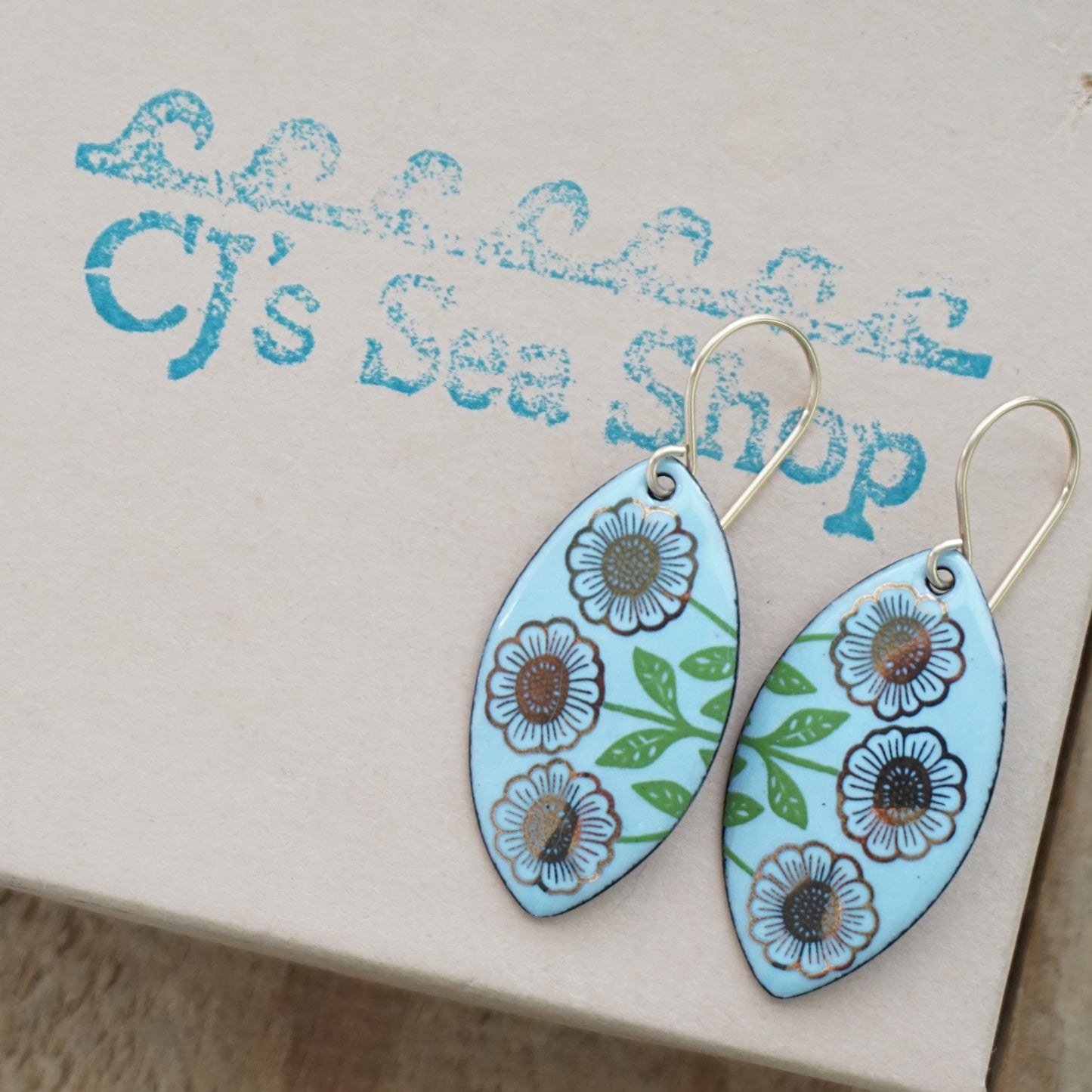 A pair of marquis-shaped drop earrings with a light blue background, decorated with gold flowers on green stems, hanging from gold hooks, displayed on a gift box