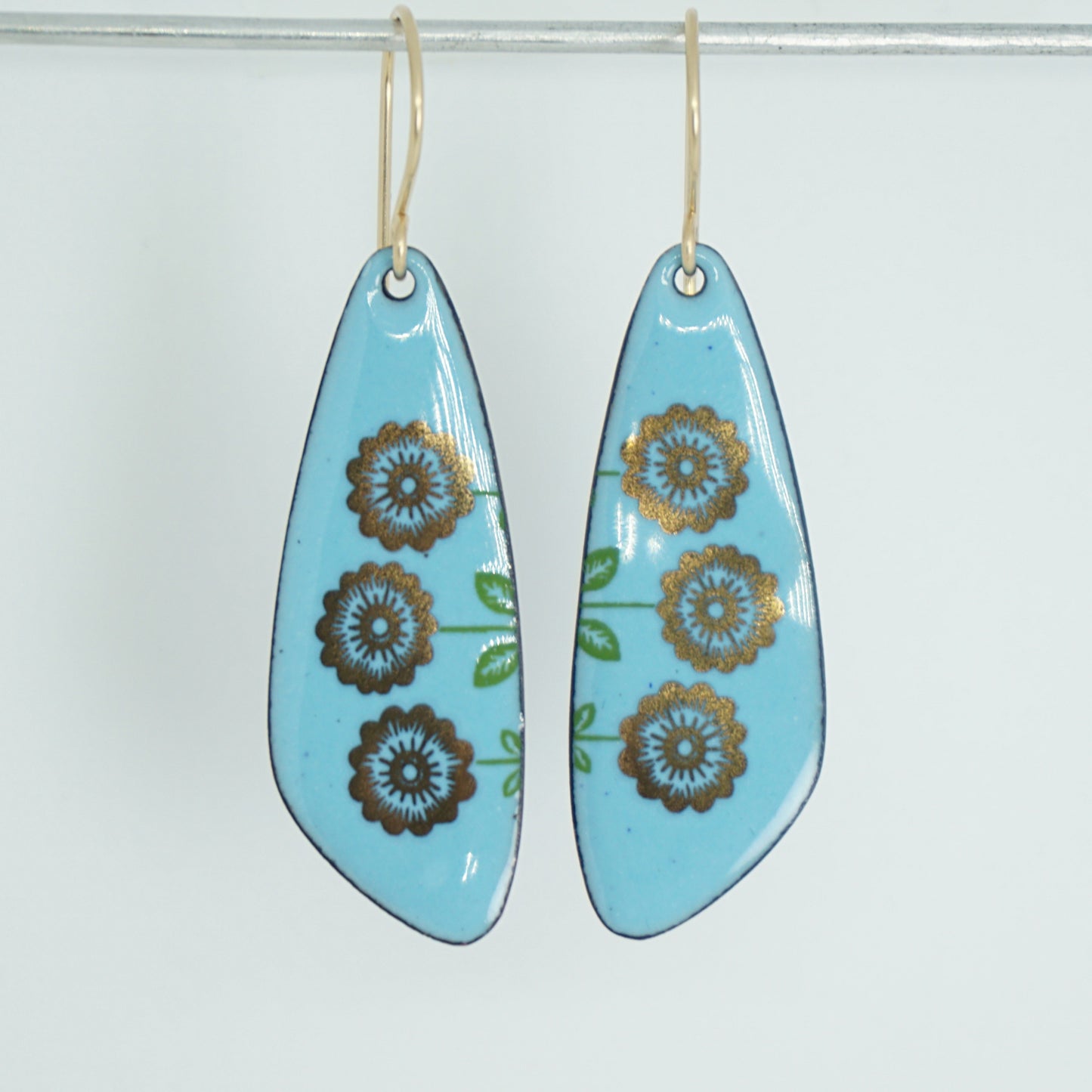 A pair of teardrop drop earrings with a light blue background, decorated with gold flowers on green stems, hanging from gold hooks, against a white background