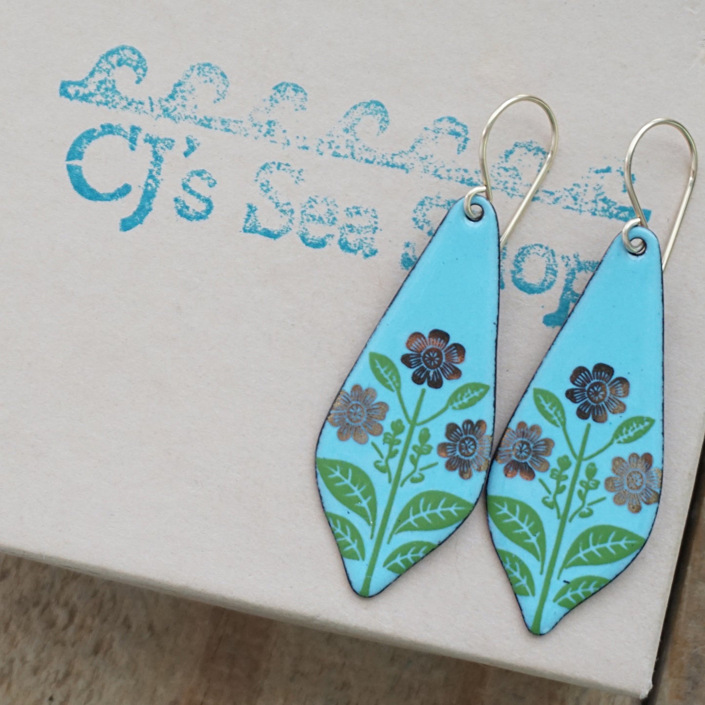 A pair of teardrop-shaped drop earrings with a light blue background, decorated with gold flowers on green stems, hanging from gold hooks, displayed on a gift box