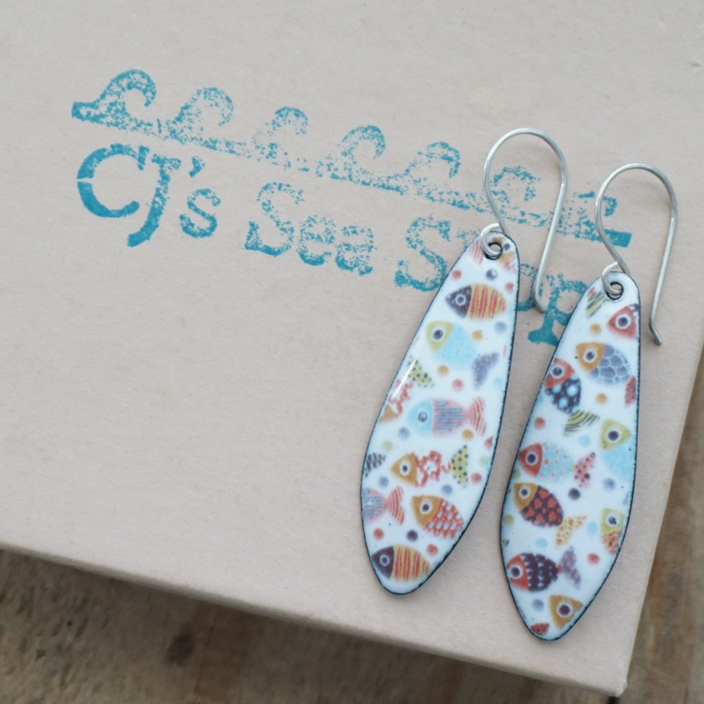 A pair of teardrop-shaped dangle earrings with colorful fish patterns on a white background, displayed on a gift box