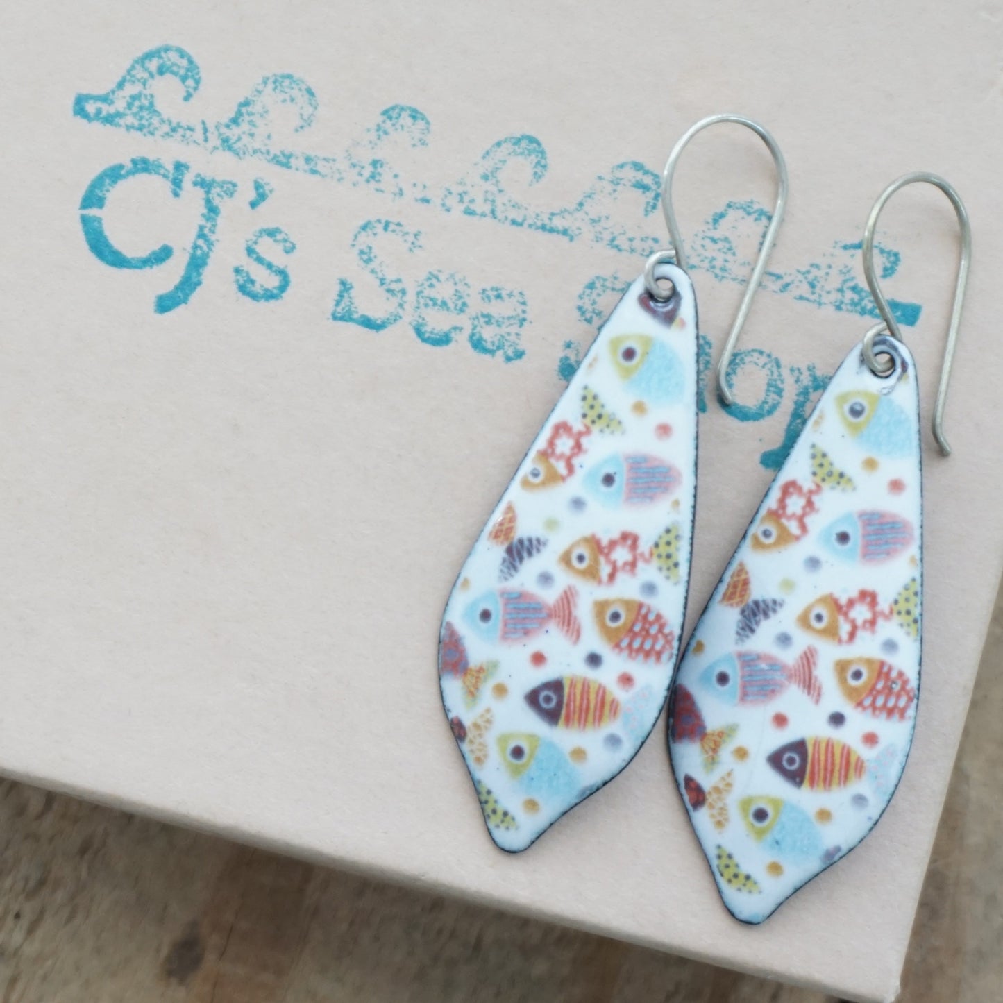 A pair of teardrop-shaped dangle earrings with a colorful fish pattern on a white background, displayed on a gift box