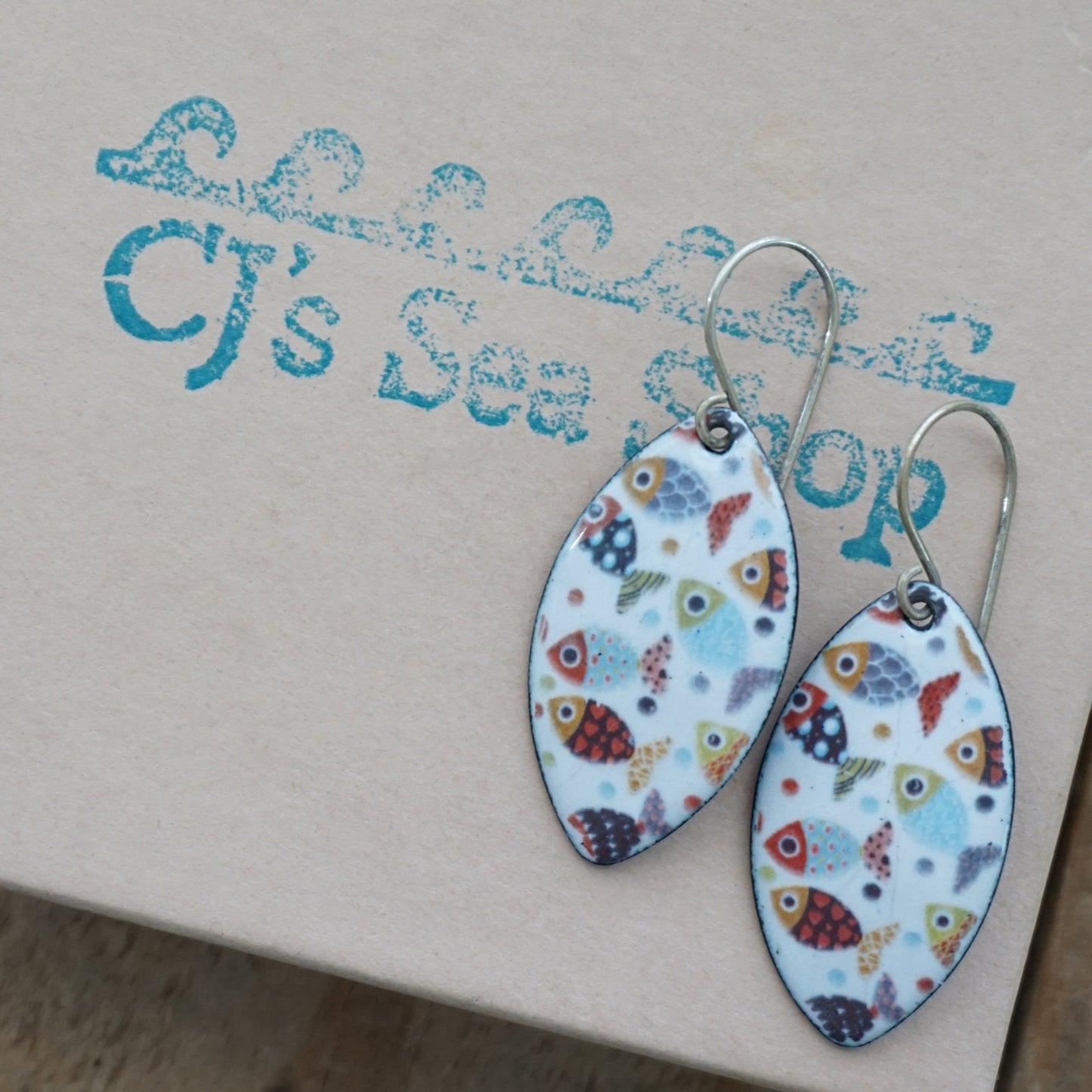 a pair of marquis shaped white enamel earrings with a colorful fish print, displayed on a gift box