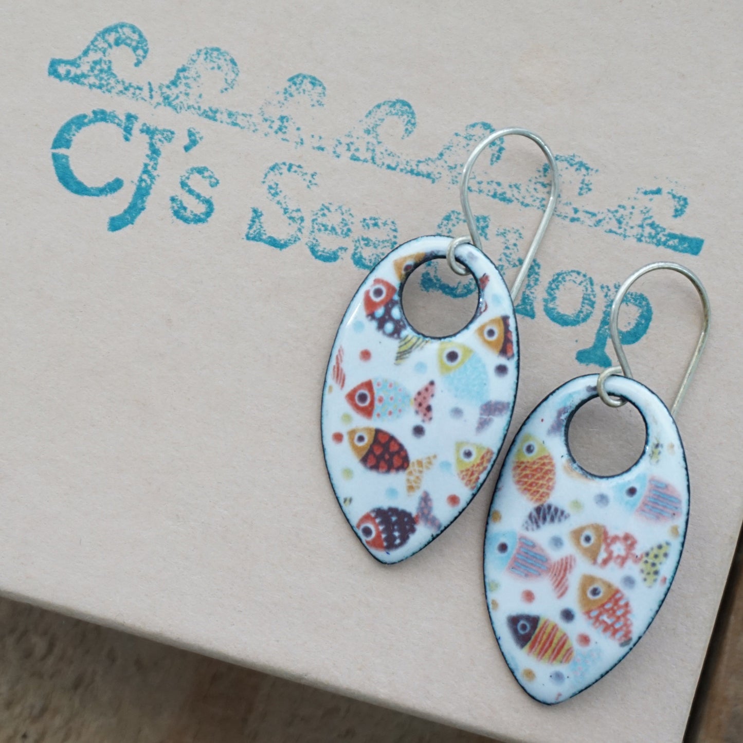 A pair of teardrop-shaped dangle earrings with a colorful fish pattern on a white background, displayed on a gift box