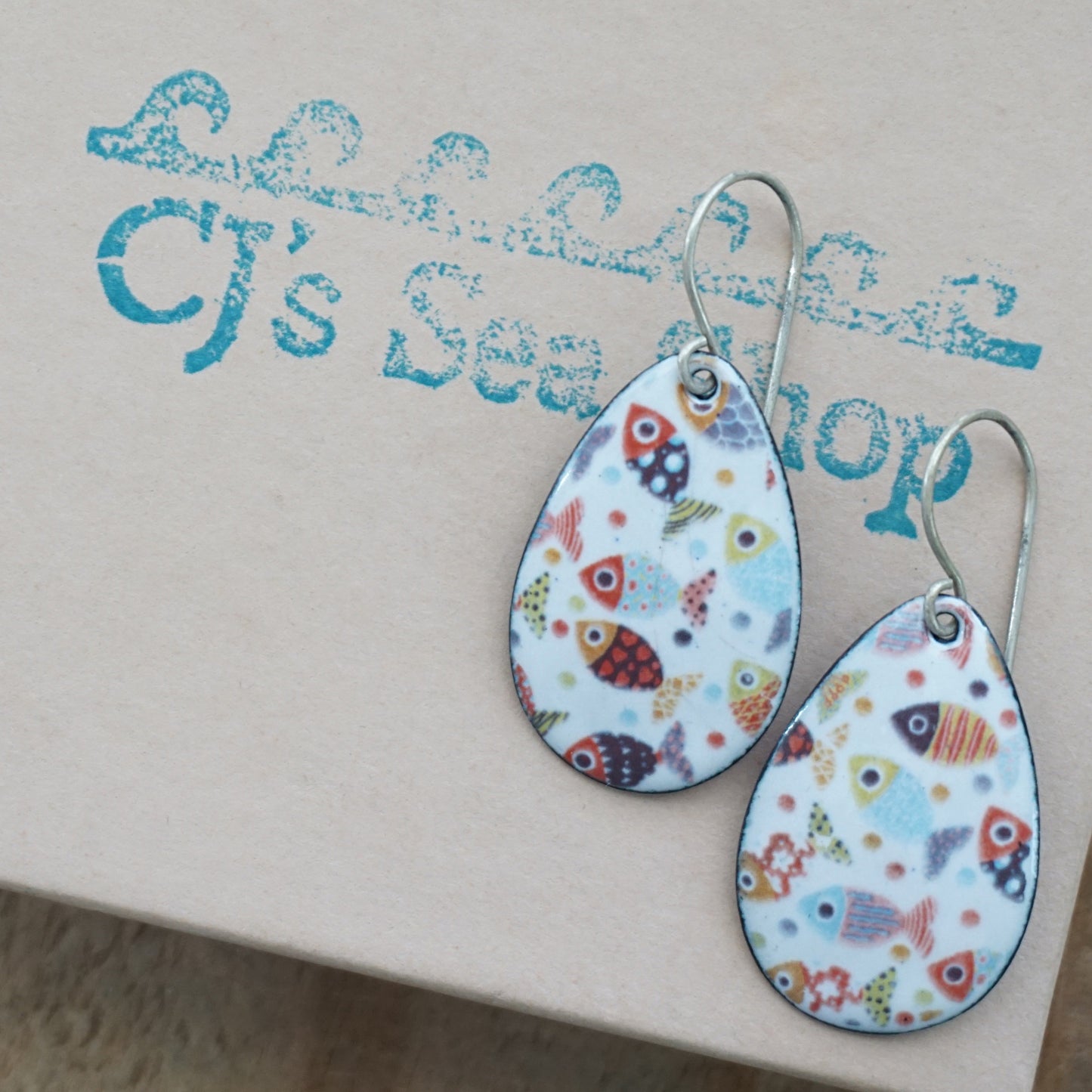 a pair of teardrop shaped white enamel earrings with a colorful fish print, displayed on a gift box