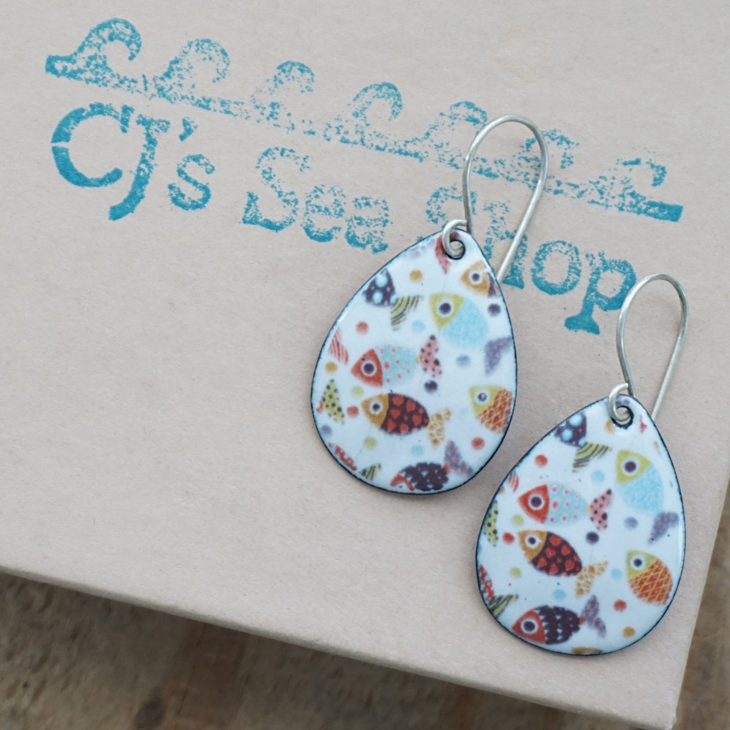 Teardrop-shaped dangle earrings with a colorful fish pattern on a white background, displayed on a gift box