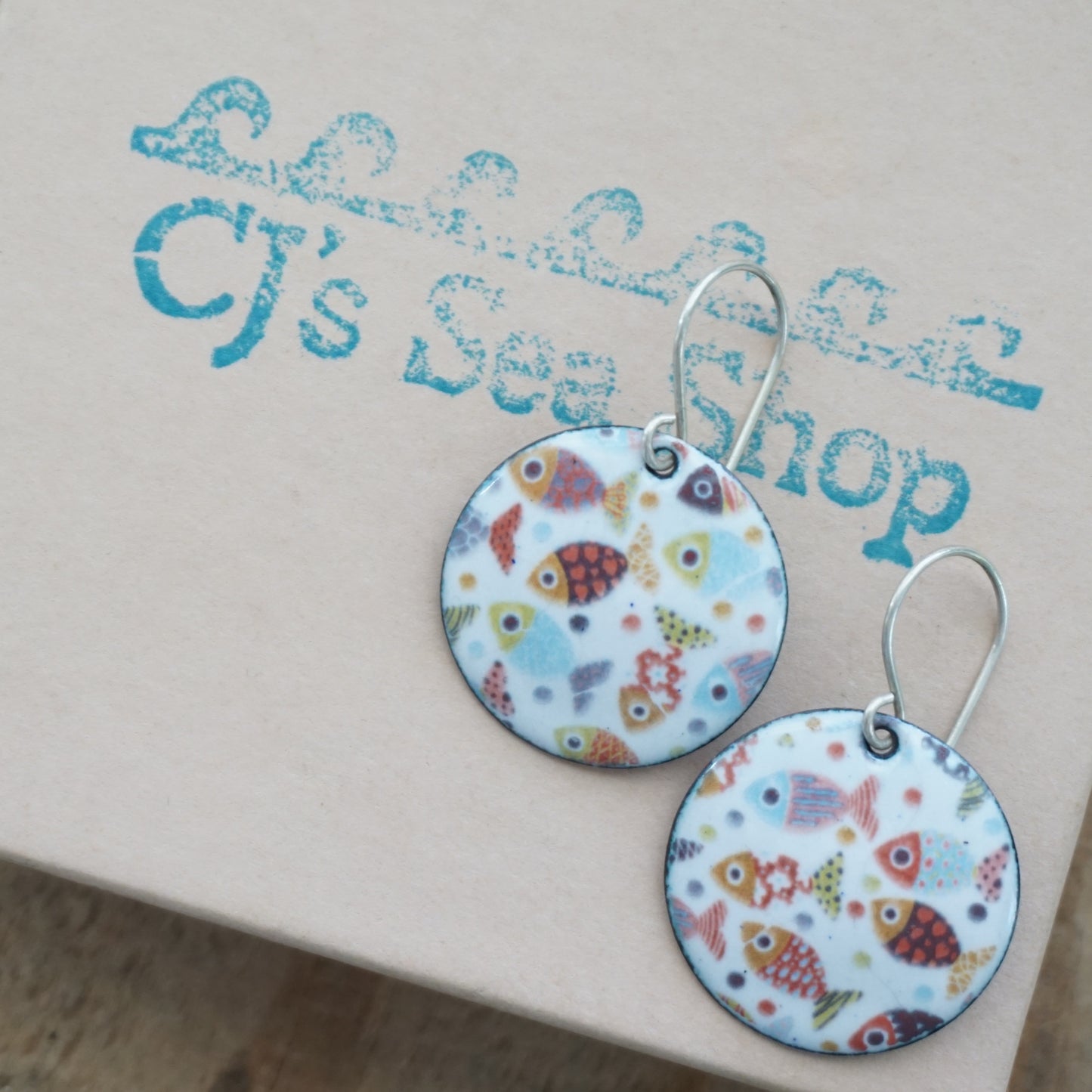 A pair of earrings with colorful fish patterns on a white background, displayed on a gift box