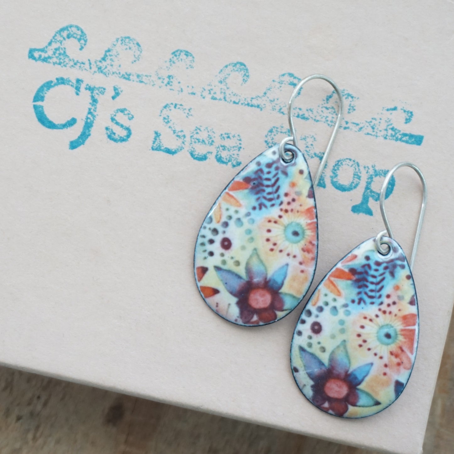 A pair of teardrop-shaped dangle earrings with a colorful floral pattern, displayed on a gift box