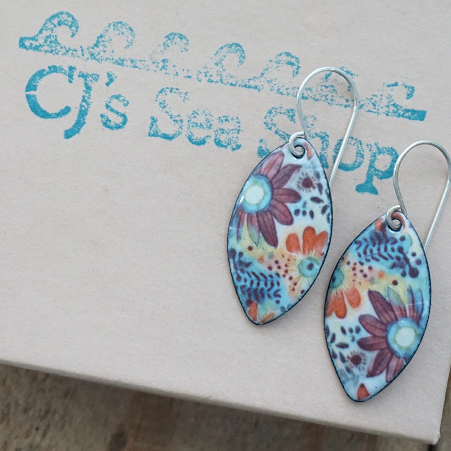 A pair of marquis-shaped dangle earrings with a colorful floral pattern on a light blue background, displayed on a gift box