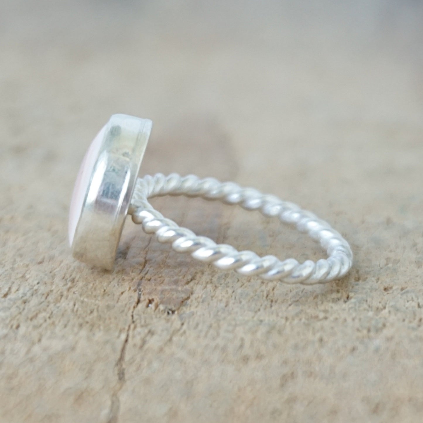 Side view of A silver ring featuring a round, smooth pale pink stone, on a wooden surface.