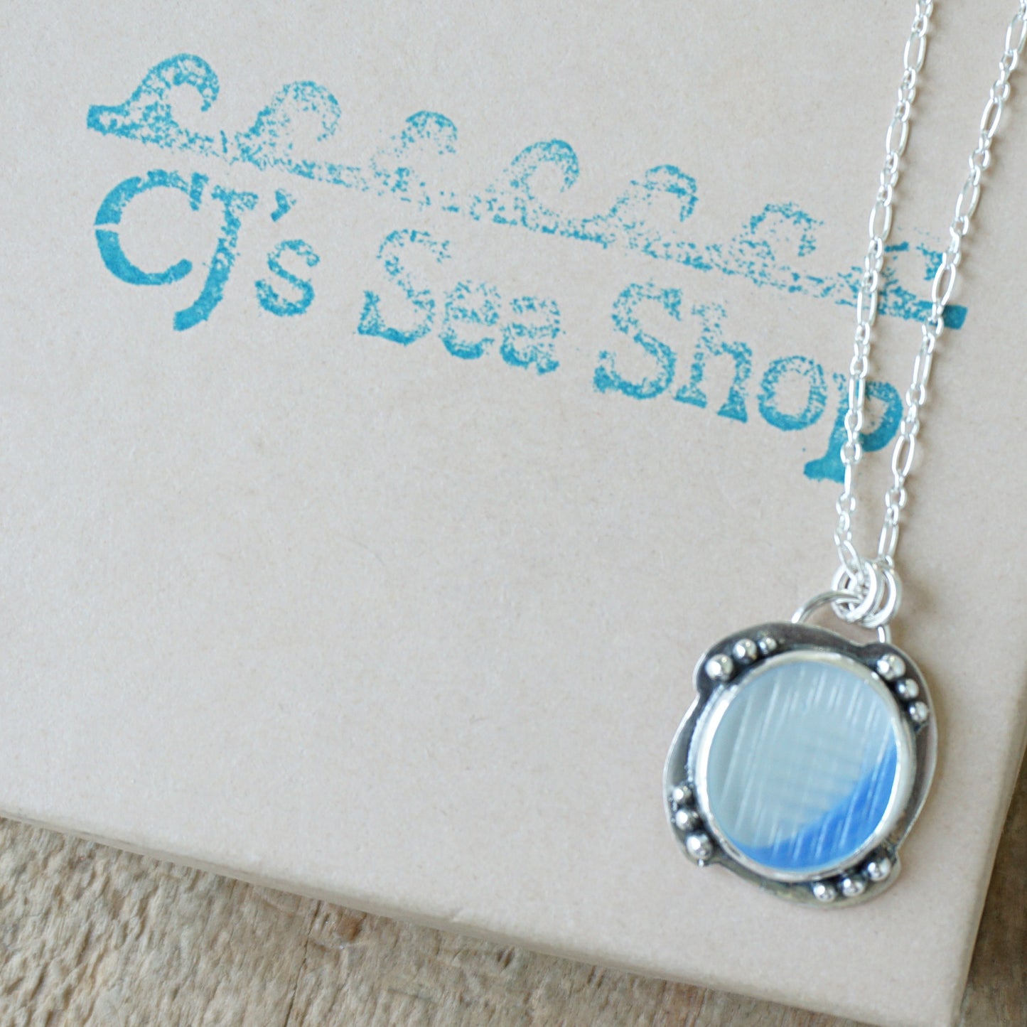 A silver pendant necklace with a round blue and clear marbled sea glass centerpiece, displayed on a gift box
