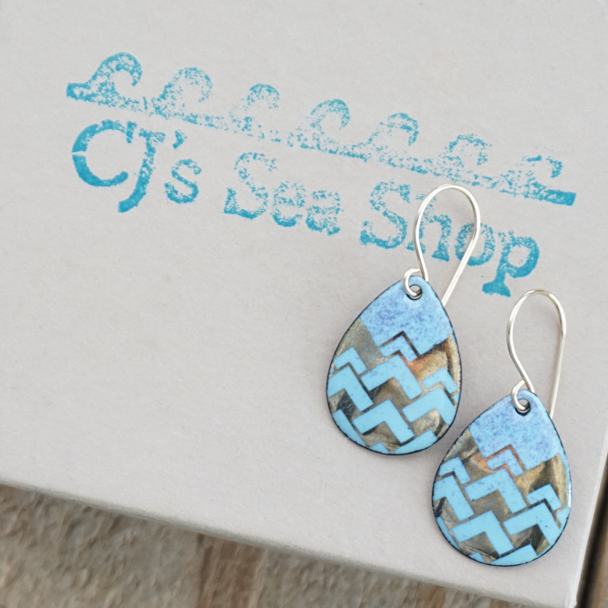 Teardrop shaped, light blue and purple enamel earrings with gold geometric designs, displayed on a gift box