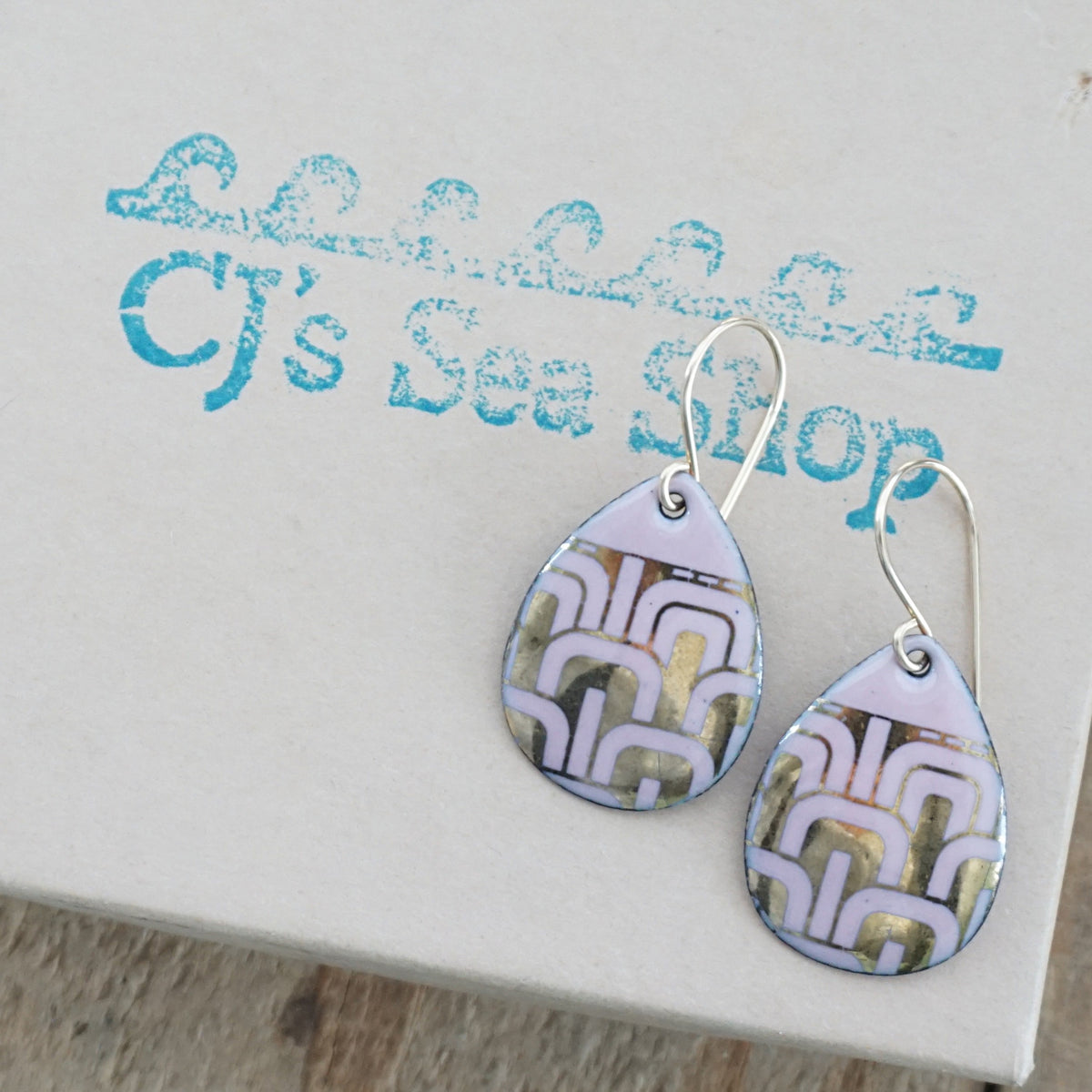 Teardrop shaped, pink enamel earrings with gold geometric designs, displayed on a gift box
