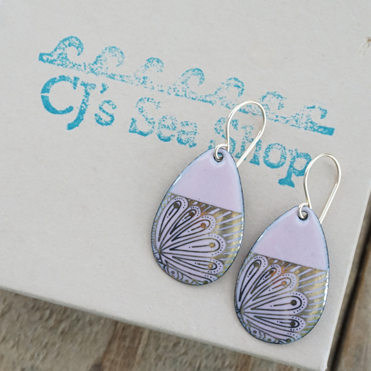 Teardrop shaped, pink enamel earrings with gold floral designs, displayed on a gift box