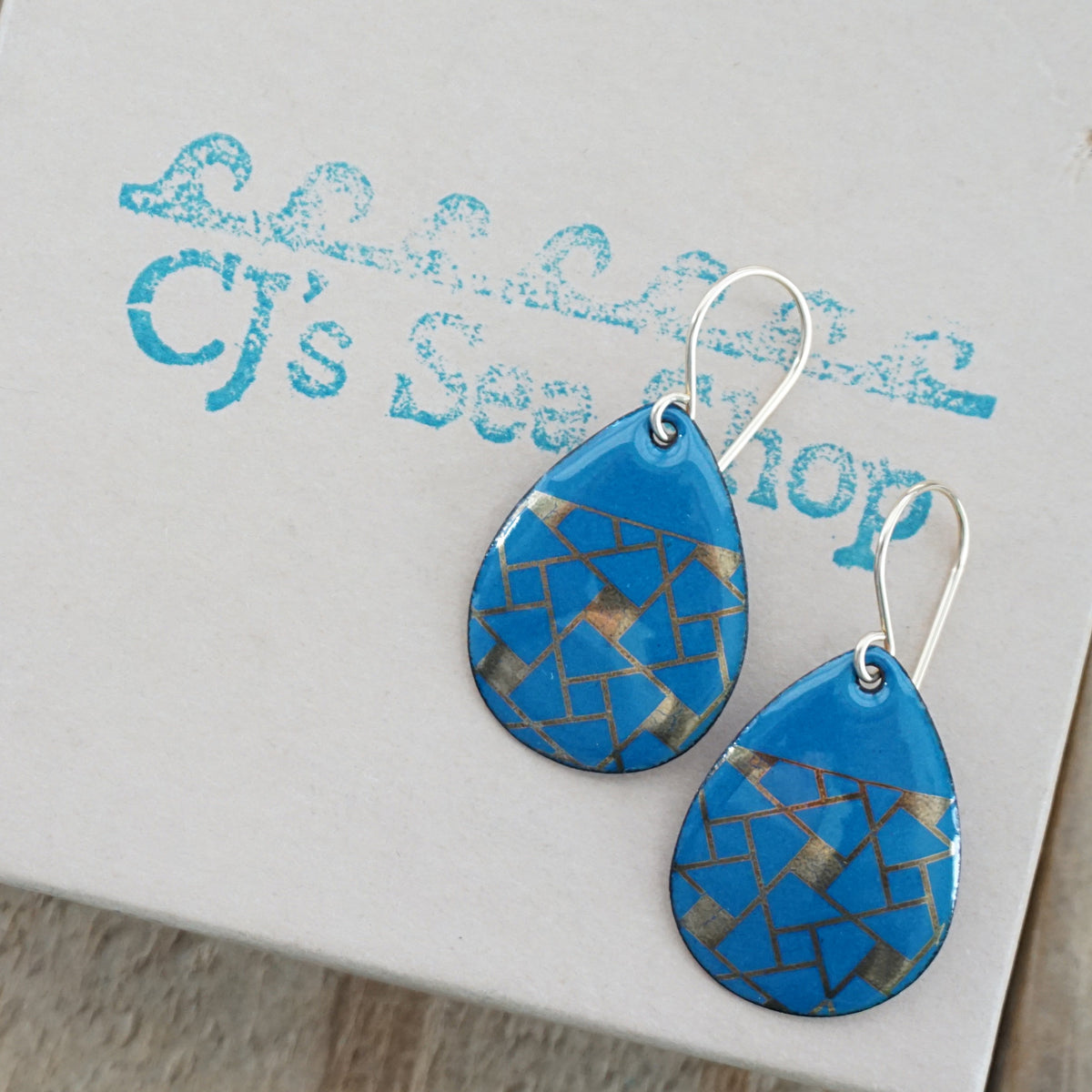 Teardrop shaped, peacock teal blue enamel earrings with gold geometric designs, displayed on a gift box