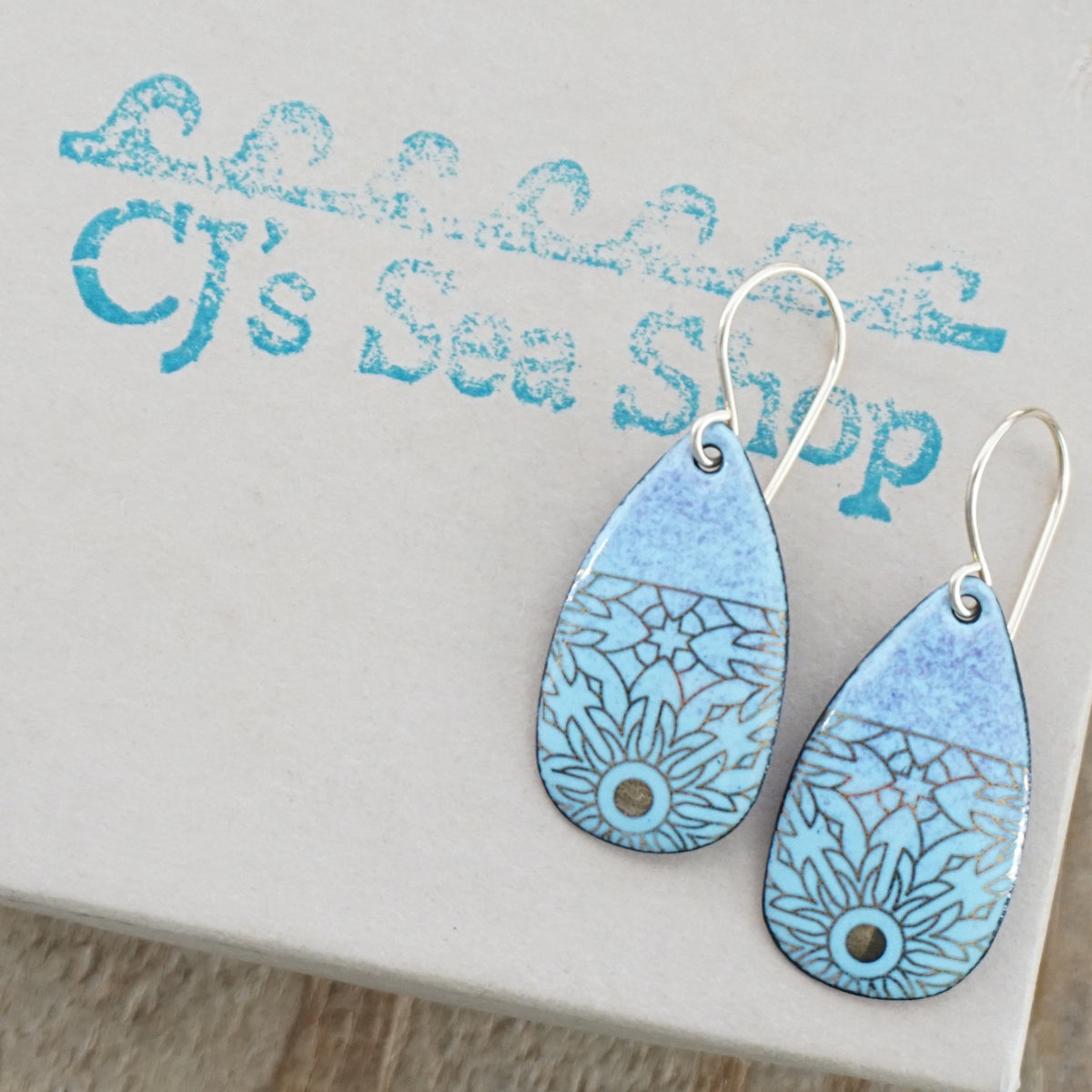 A pair of teardrop-shaped dangling earrings with a light blue and lavender-purple gradient, decorated with gold floral pattern, hanging from gold hooks, displayed on a gift box