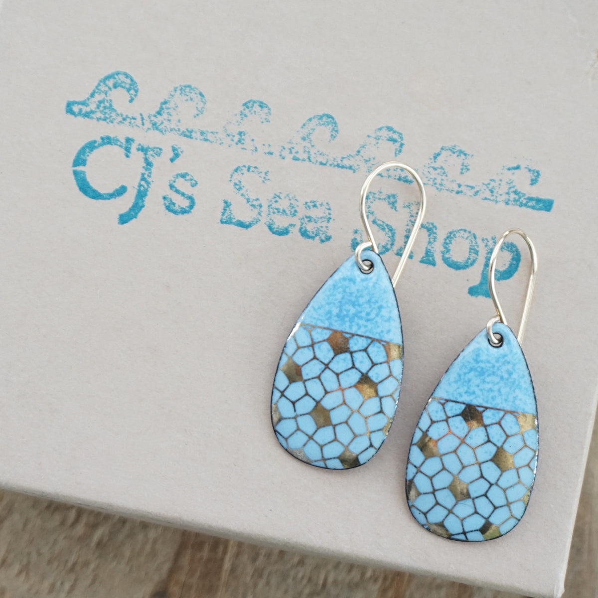 Teardrop shaped light blue enamel earrings with a gold geometric pattern, displayed on a gift box