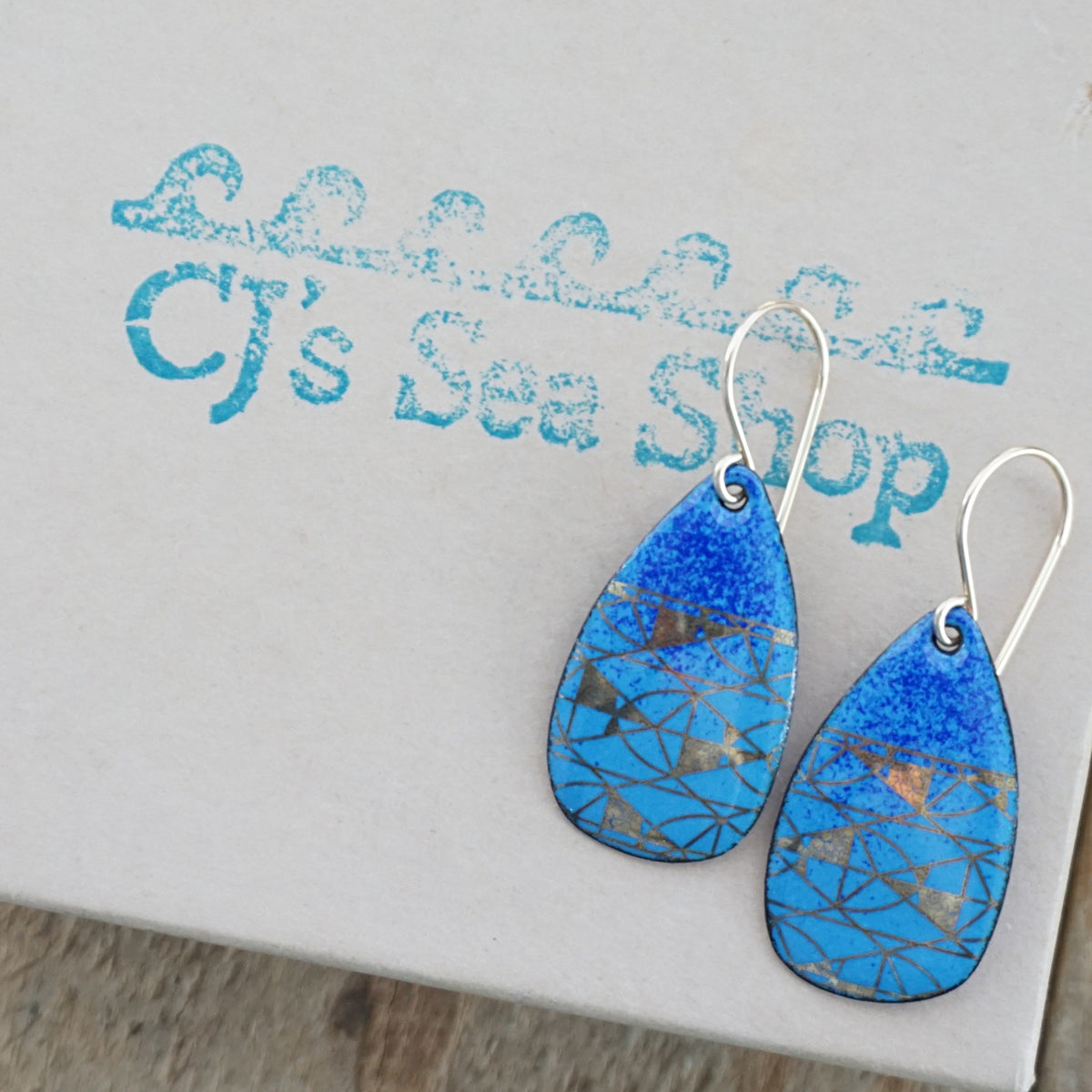 A pair of teardrop-shaped blue enamel earrings with geometric gold foil patterns and gold-tone wire hooks, displayed on a gift box
