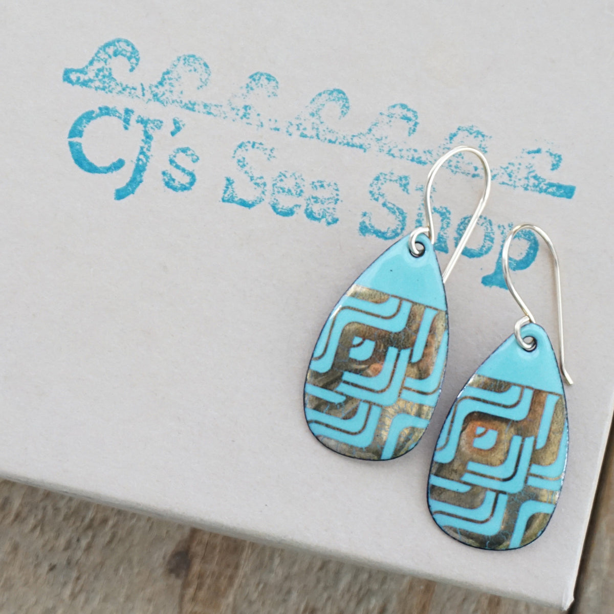 A pair of teardrop-shaped seafoam green enamel earrings with geometric gold foil patterns and gold-tone wire hooks, displayed on a gift box