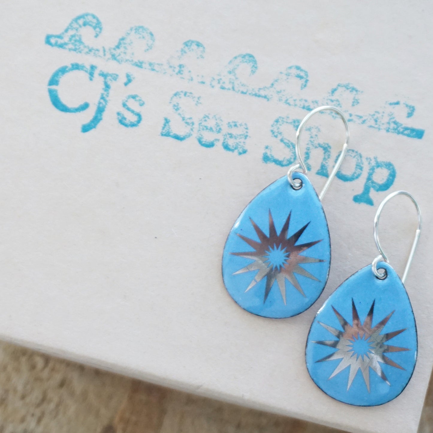 A pair of blue enamel teardrop earrings with silver snowflake designs, on a gift box