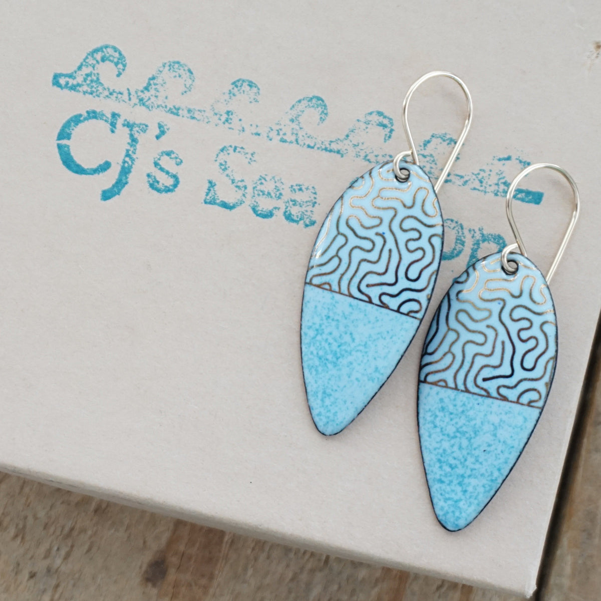 a pair of light blue enamel earrings with gold designs, displayed on a gift box