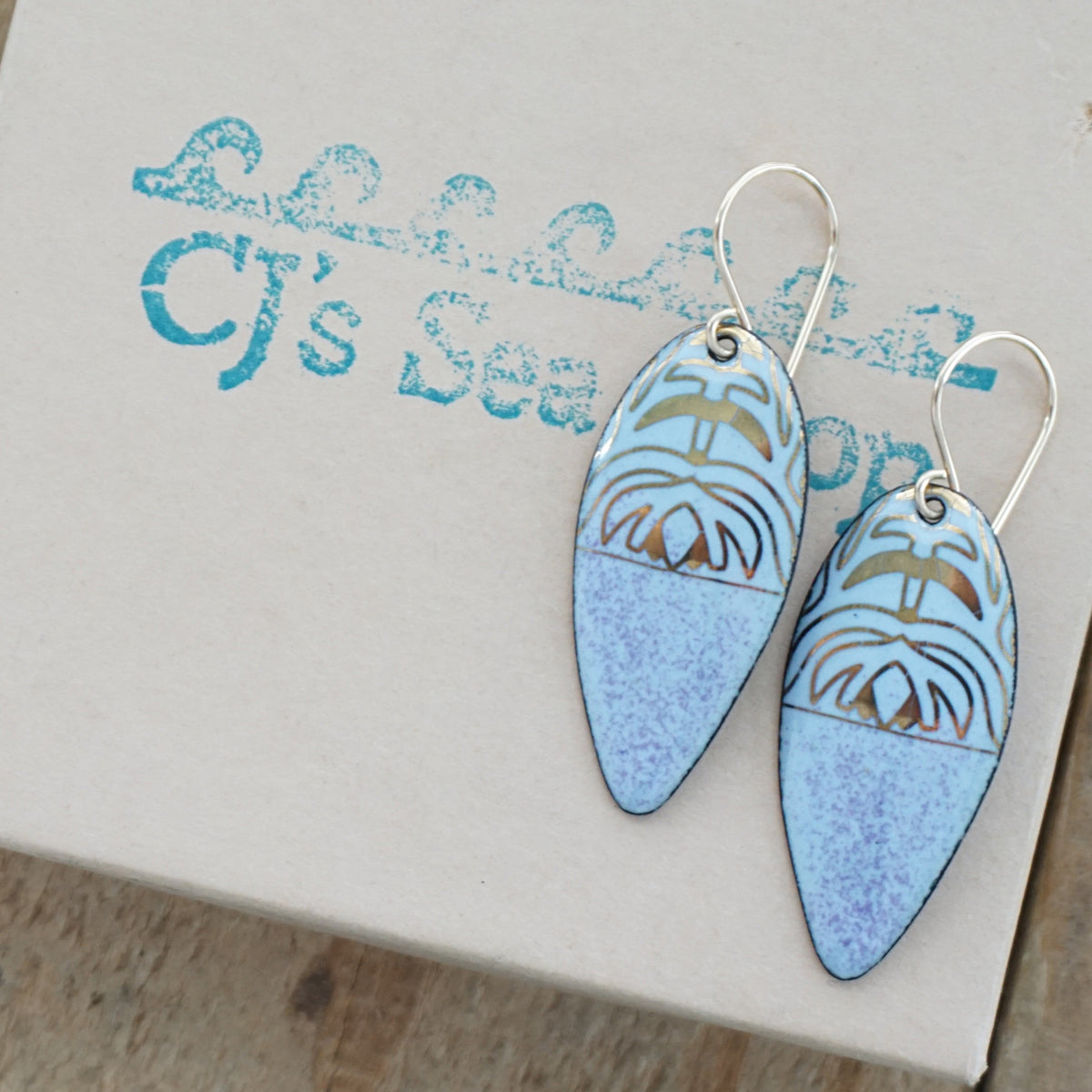 a pair of light purple enamel earrings with gold designs, displayed on a gift box