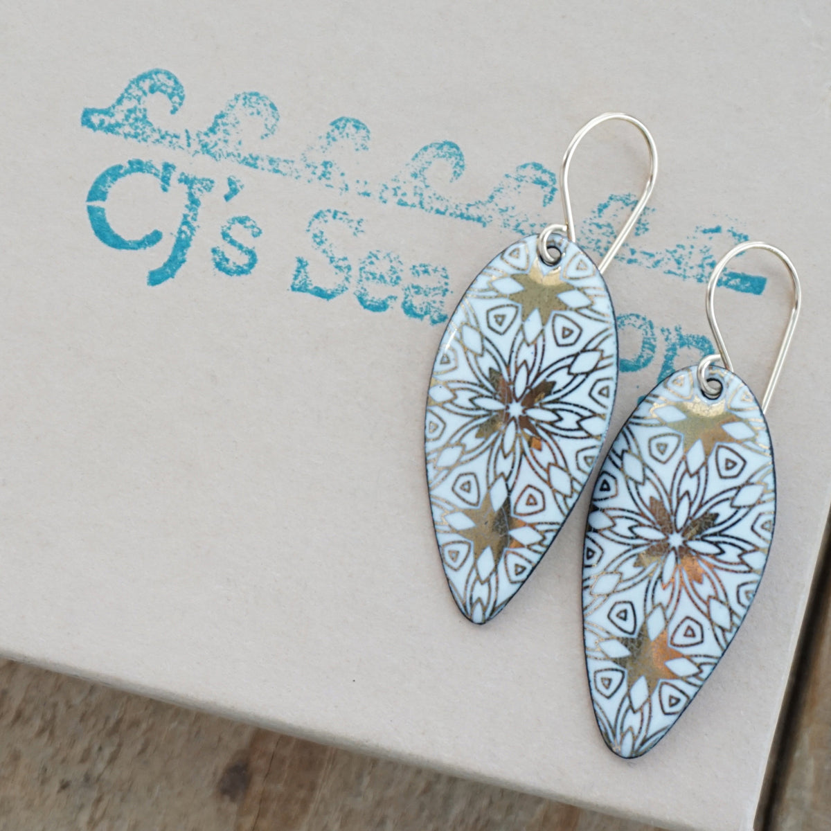 a pair of white enamel earrings with gold designs, displayed on a gift box
