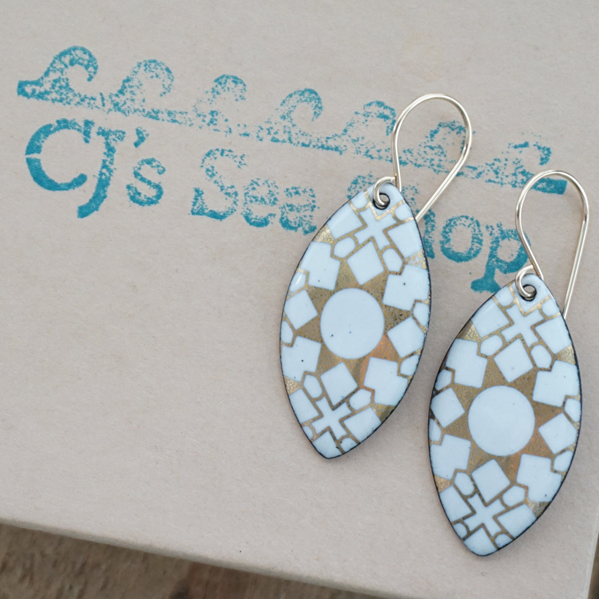 a pair of white enamel earrings with gold designs, displayed on a gift box