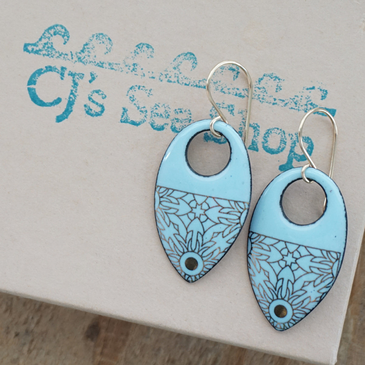 a pair of light blue enamel earrings with gold designs, displayed on a gift box