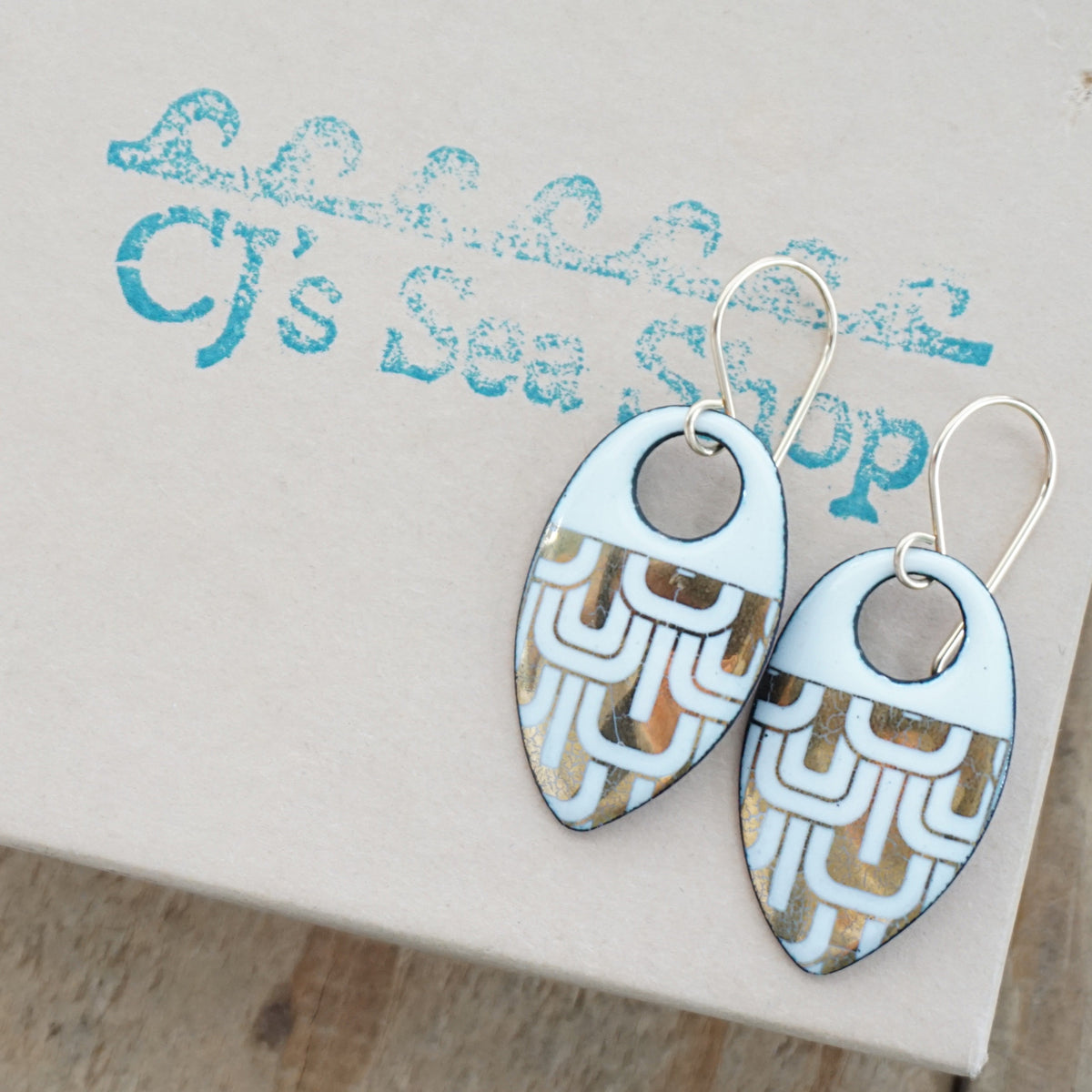 a pair of white enamel earrings with gold designs, displayed on a gift box
