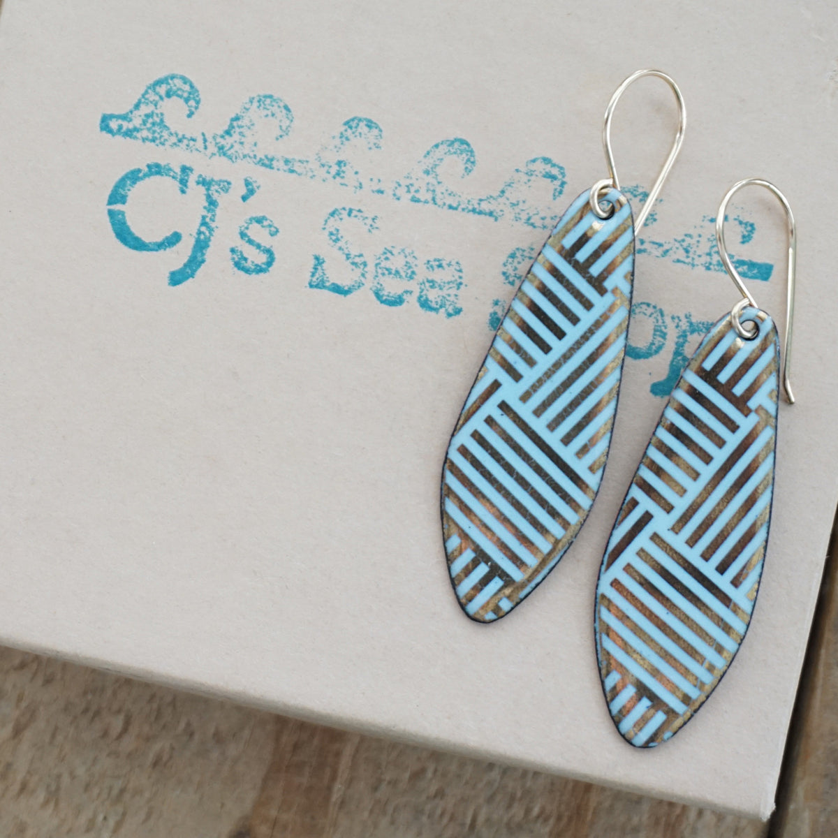a pair of light blue enamel earrings with gold designs, displayed on a gift box