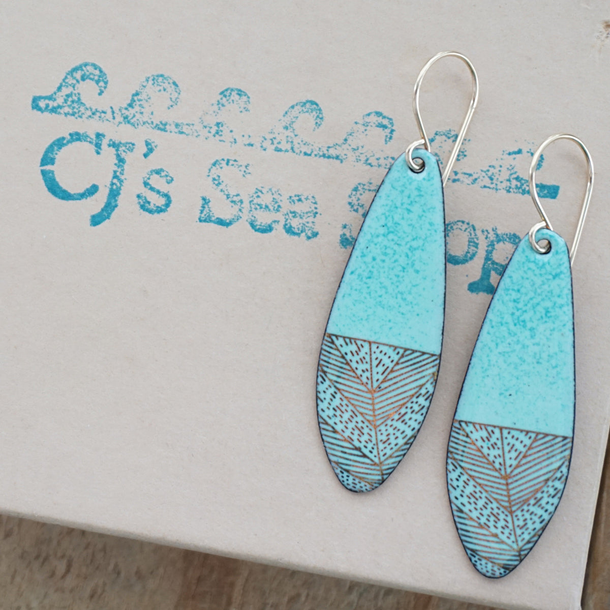 a pair of seafoam green enamel earrings with gold designs, displayed on a gift box