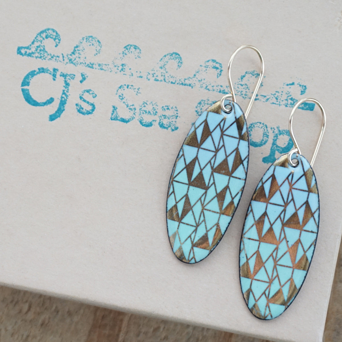 a pair of light blue and green enamel earrings with gold designs, displayed on a gift box