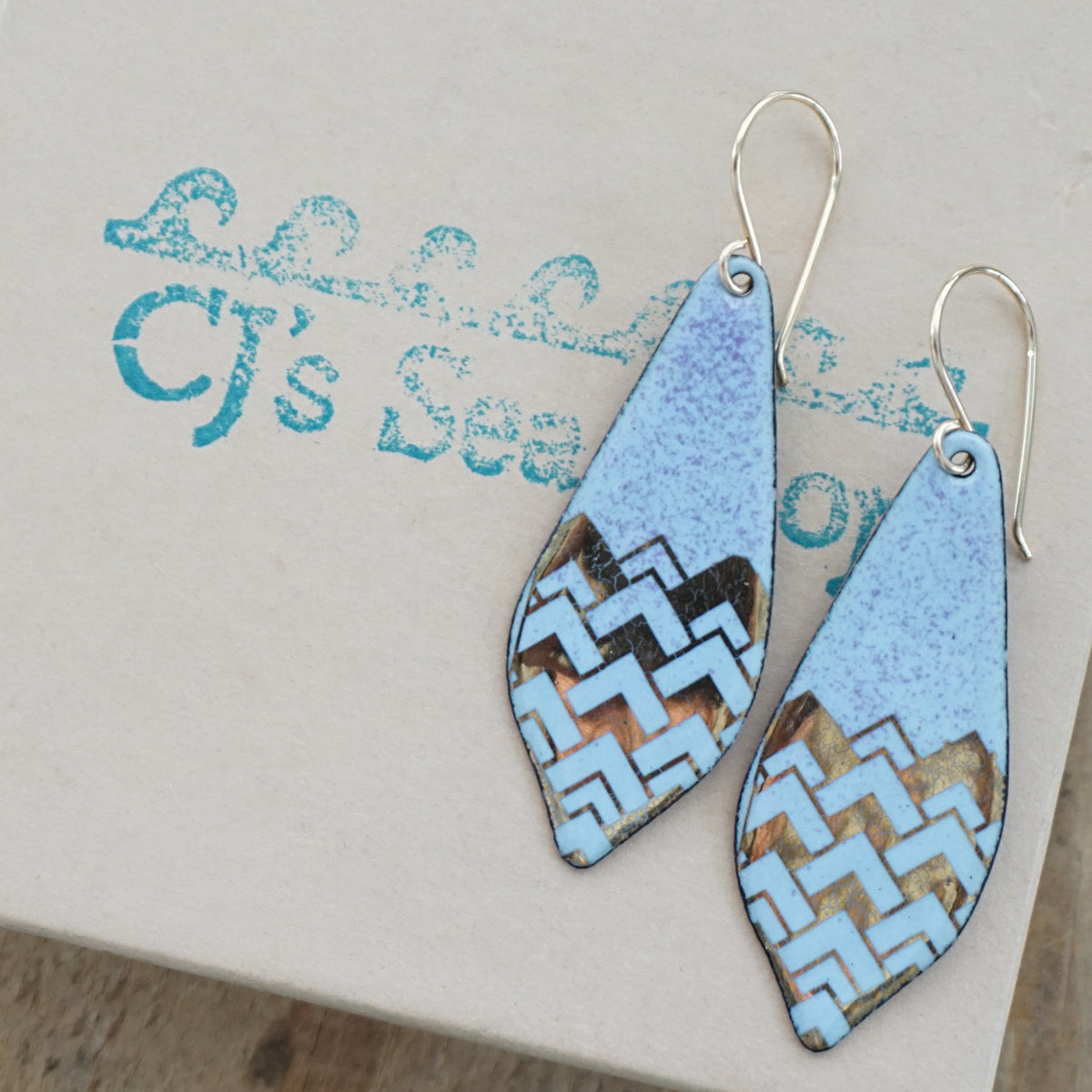 a pair of light purple enamel earrings with gold designs, displayed on a gift box
