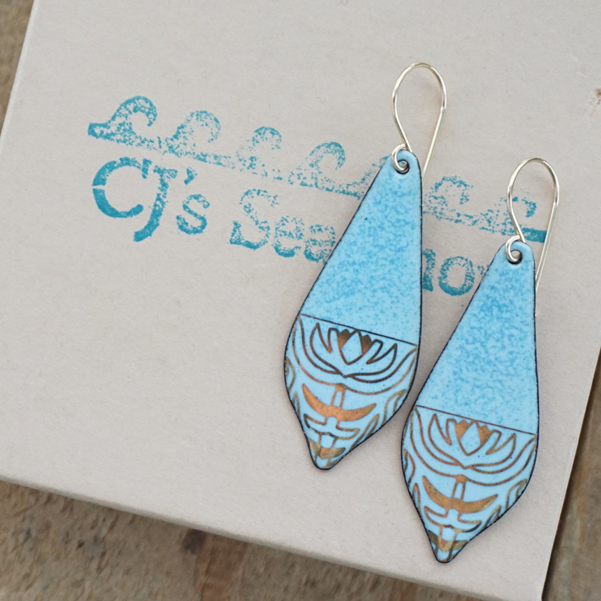 a pair of light blue enamel earrings with gold designs, displayed on a gift box